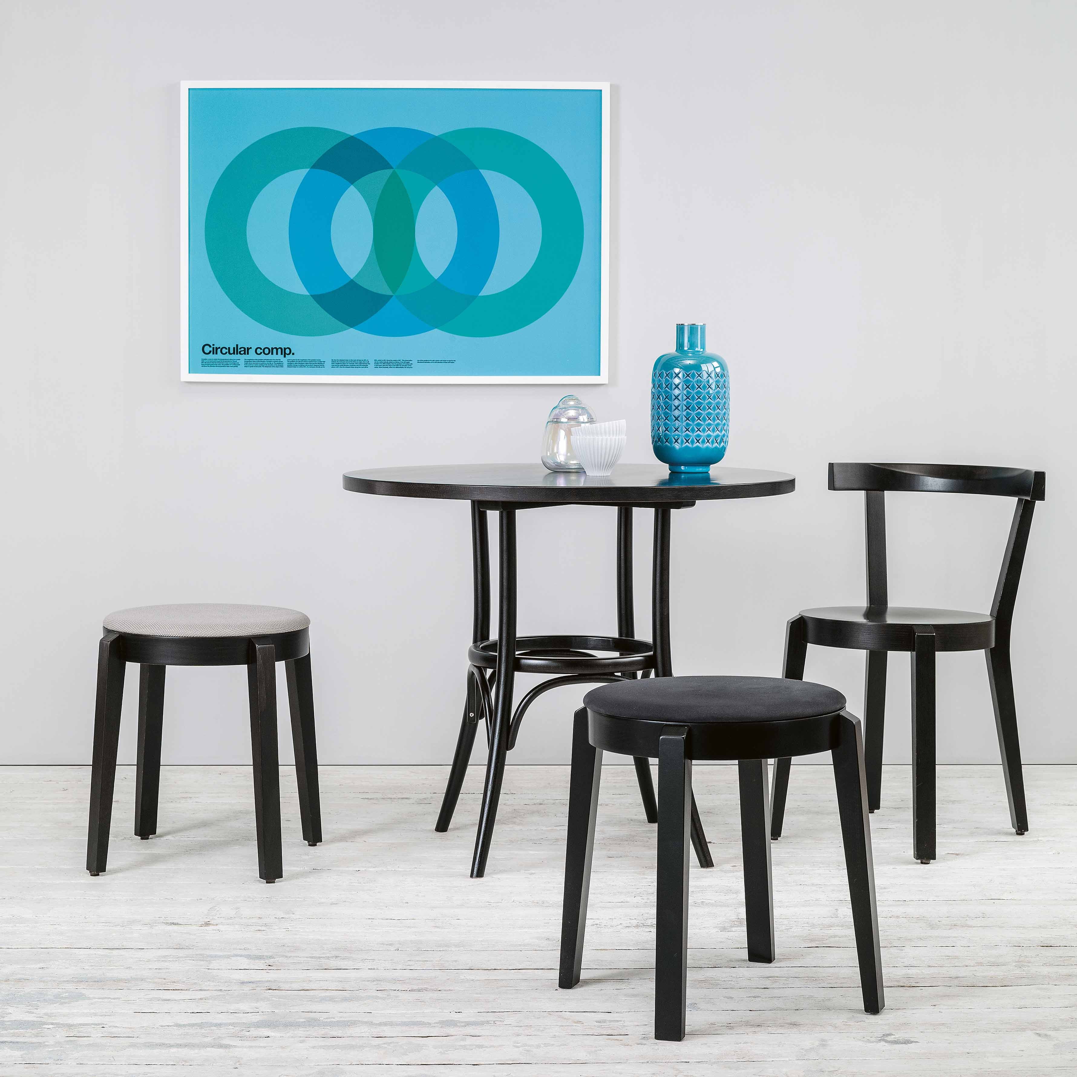 A minimalist dining area showcases a round black table with three TON Stool Puntons. A turquoise patterned vase and a small glass rest on the table, while an abstract blue circular art piece adorns the gray walls above light wood floors, exuding universal use and style.