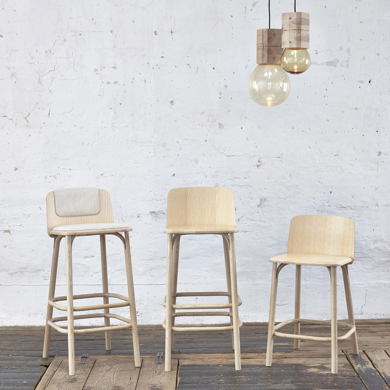 Three light TON Barstool Split pieces, showcasing Arik Levys craftsmanship, are set on a rustic wooden floor against a textured white wall. Above, two wooden pendant lights with clear bulbs artfully illuminate the space.