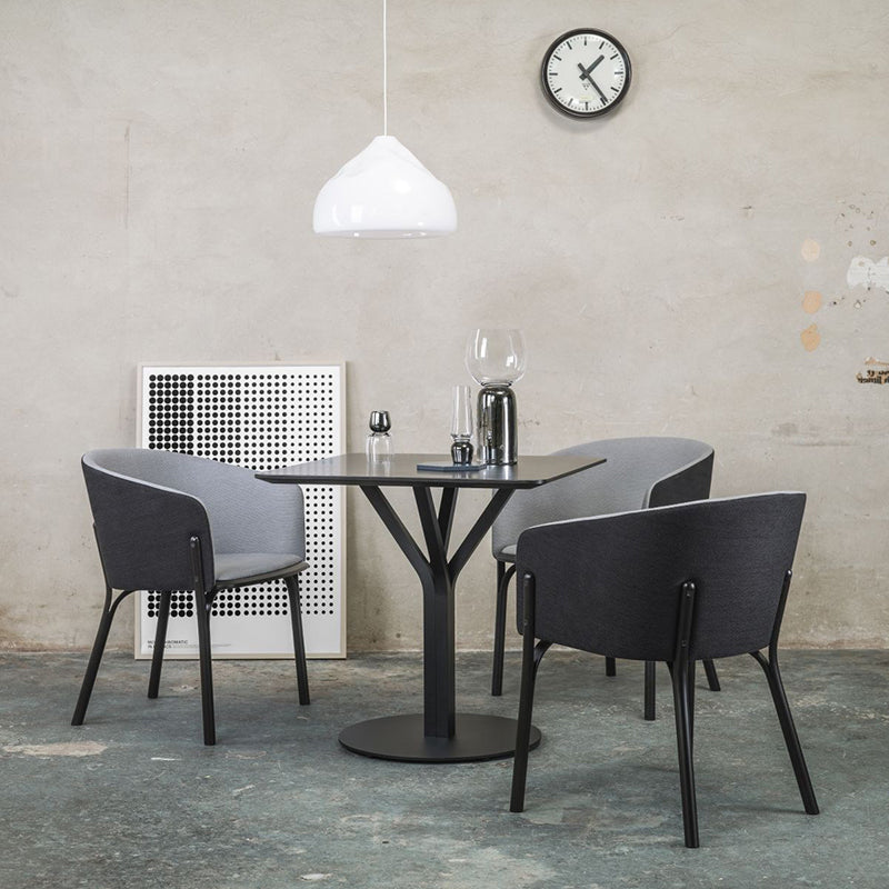 A modern dining room boasts a round black table with three TON Armchair Splits in gray, upholstered for comfort. Set on a concrete floor, the space features a white pendant light casting warmth and is accented by a white ventilation panel and wall clock on a beige wall, exuding classic TONDesigner elegance.