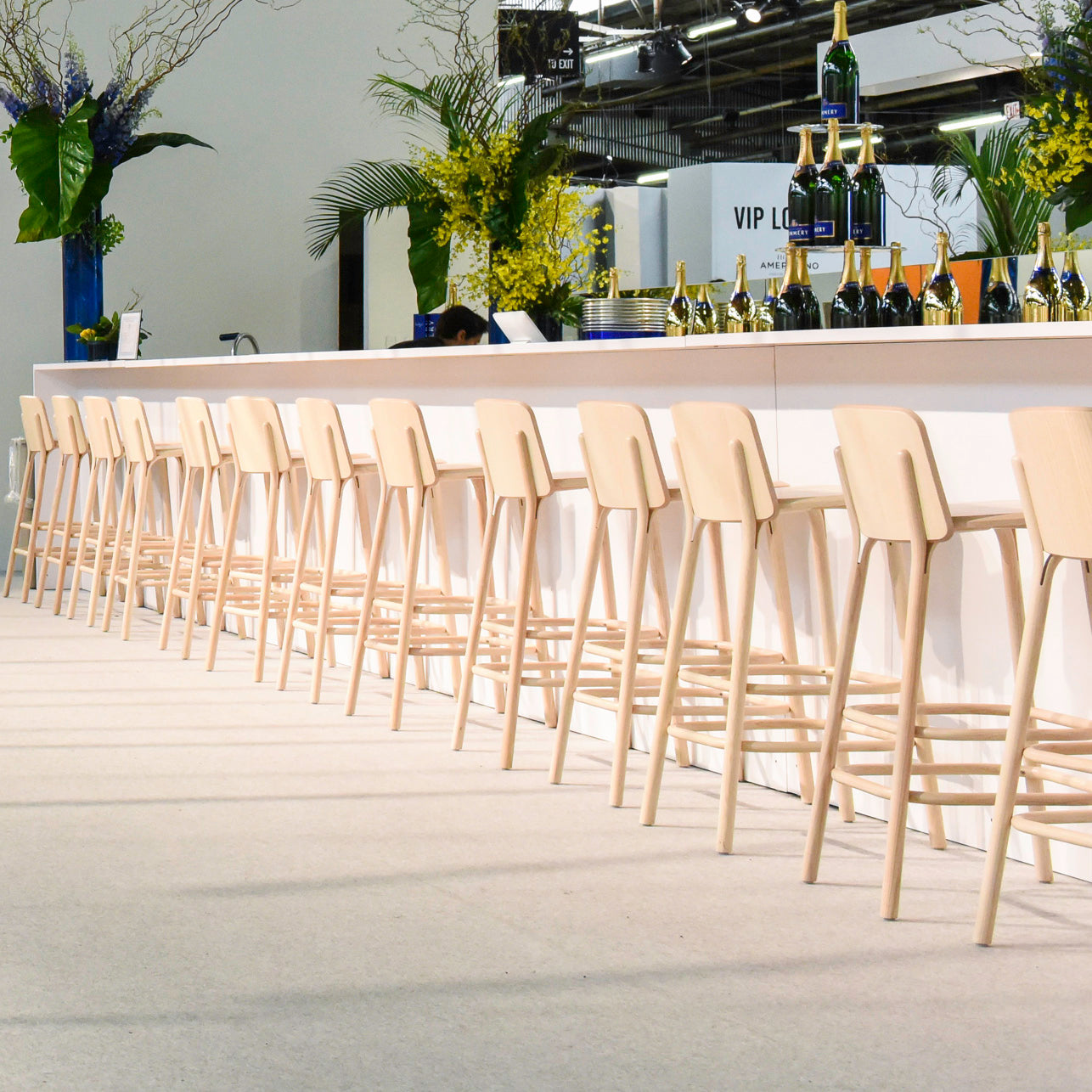 A modern bar showcases chic TON Barstool Split seats aligned at the front. Champagne bottles and flutes grace the bar, harmonized with yellow flowers and large leaves in the decor. A visible VIP Lounge sign enhances exclusivity in this stylish space inspired by Arik Levy.