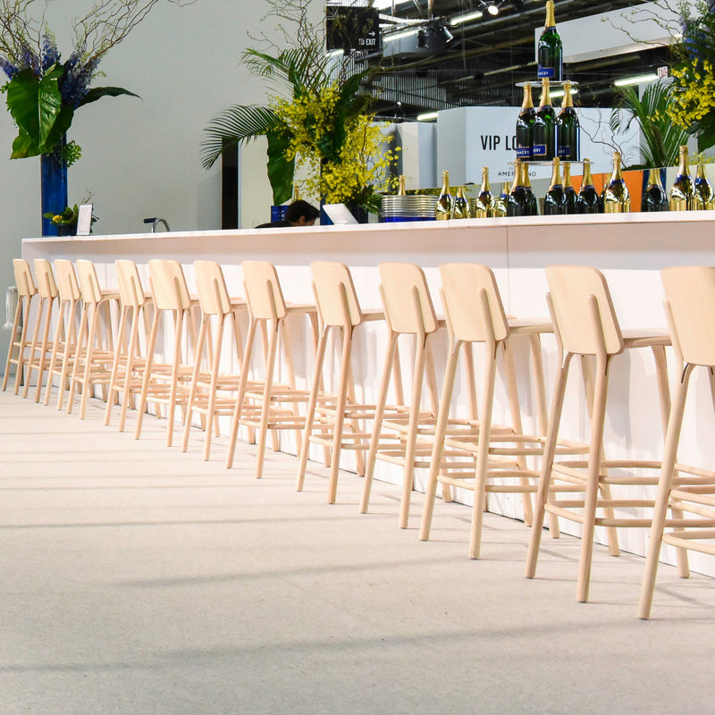 A modern bar showcases chic TON Barstool Split seats aligned at the front. Champagne bottles and flutes grace the bar, harmonized with yellow flowers and large leaves in the decor. A visible VIP Lounge sign enhances exclusivity in this stylish space inspired by Arik Levy.