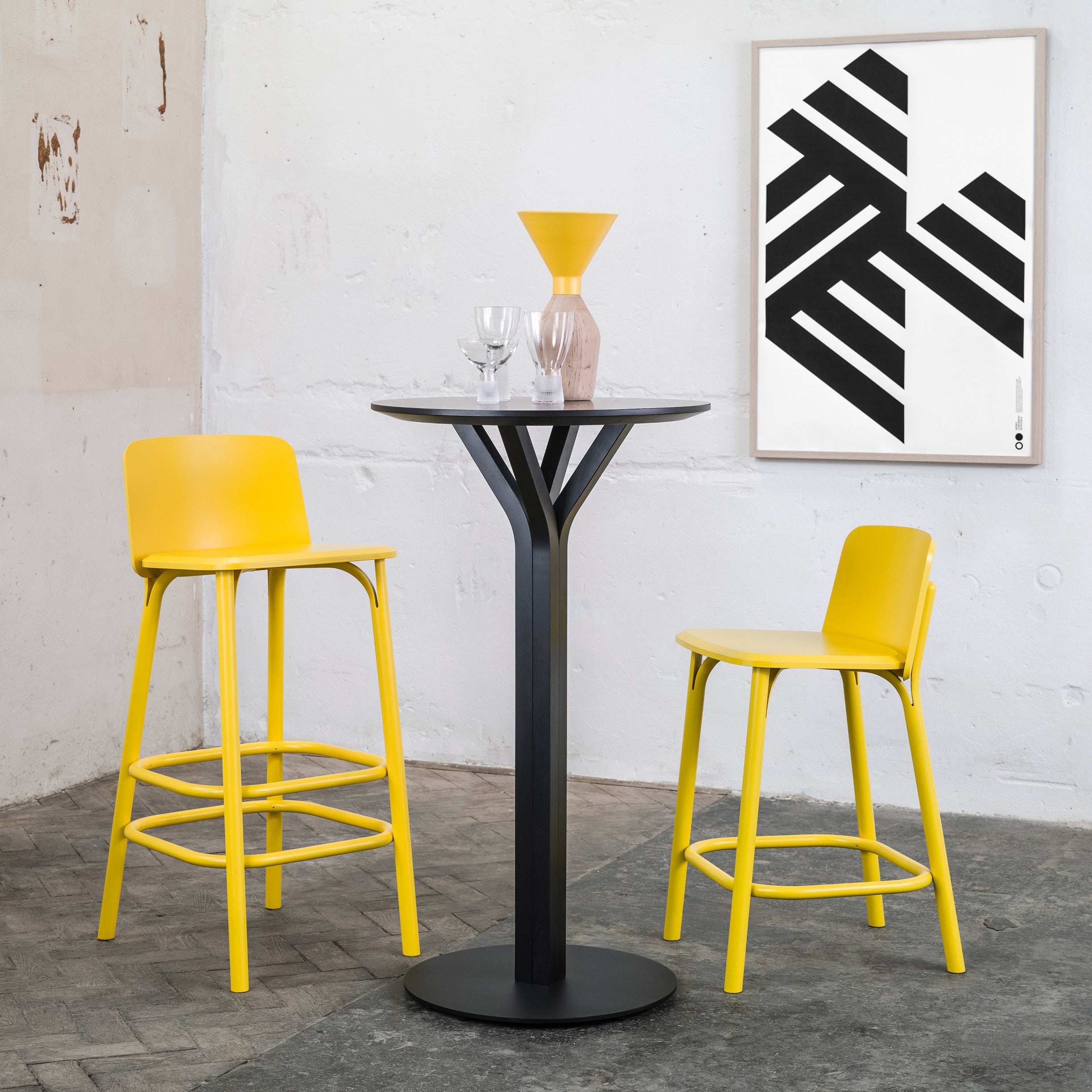 In a modern cafe, a tall black table with two vibrant yellow Barstool Split - Sale by TON stands out. The table displays a matching yellow vase and two glasses, while black and white geometric artwork decorates the white wall. Rustic-style tiles complete the chic ambiance.
