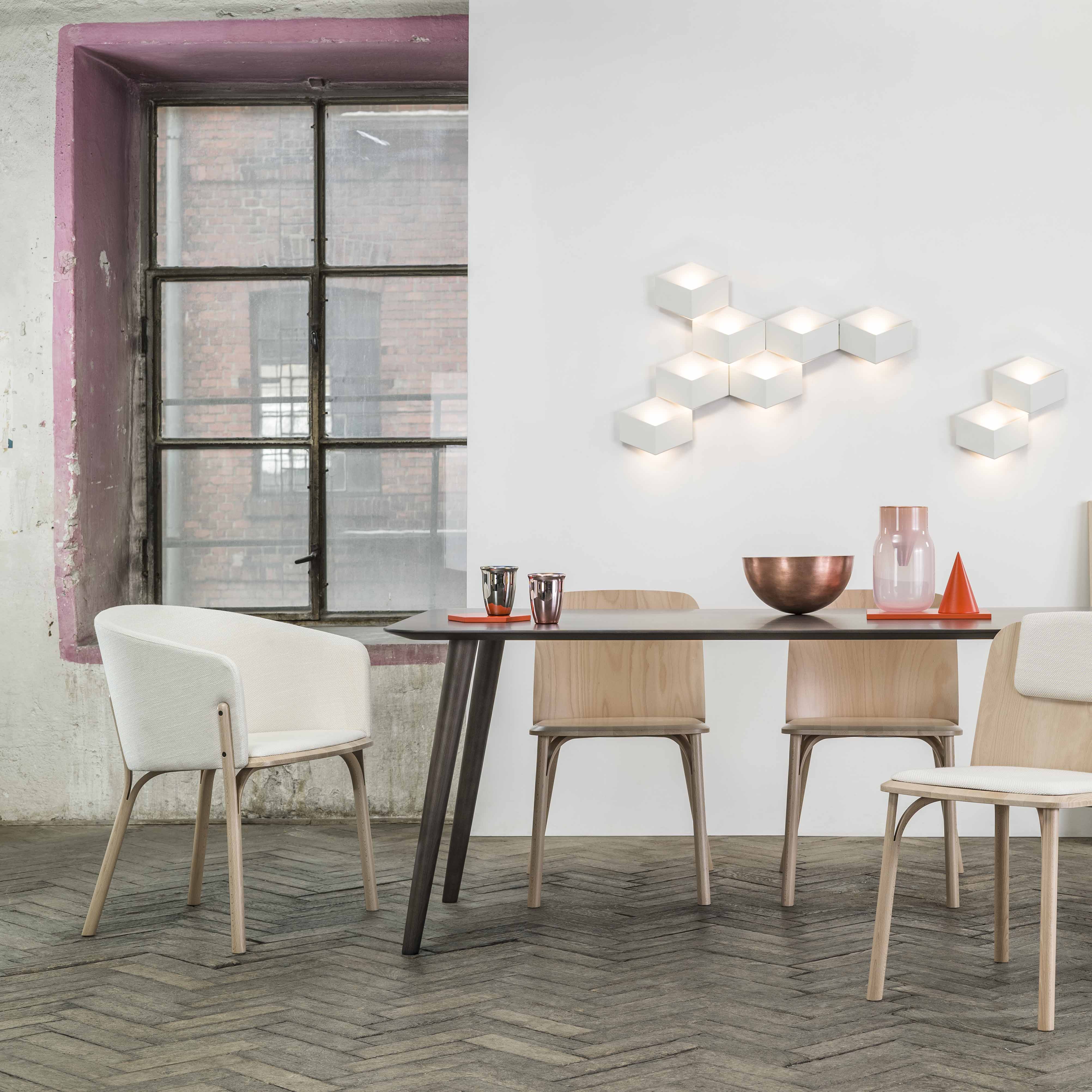 A modern dining room features a black table and TON Lounge Chair Split with wooden craftsmanship. White cushioned seats offer comfort, while a copper bowl and pitcher decorate the table. Hexagonal wall lights add charm, alongside a large pink-framed window and herringbone-patterned floor.