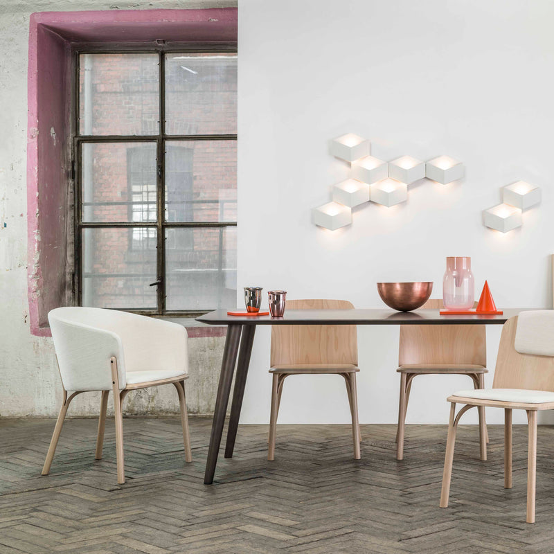 A modern dining room features a black table and TON Lounge Chair Split with wooden craftsmanship. White cushioned seats offer comfort, while a copper bowl and pitcher decorate the table. Hexagonal wall lights add charm, alongside a large pink-framed window and herringbone-patterned floor.