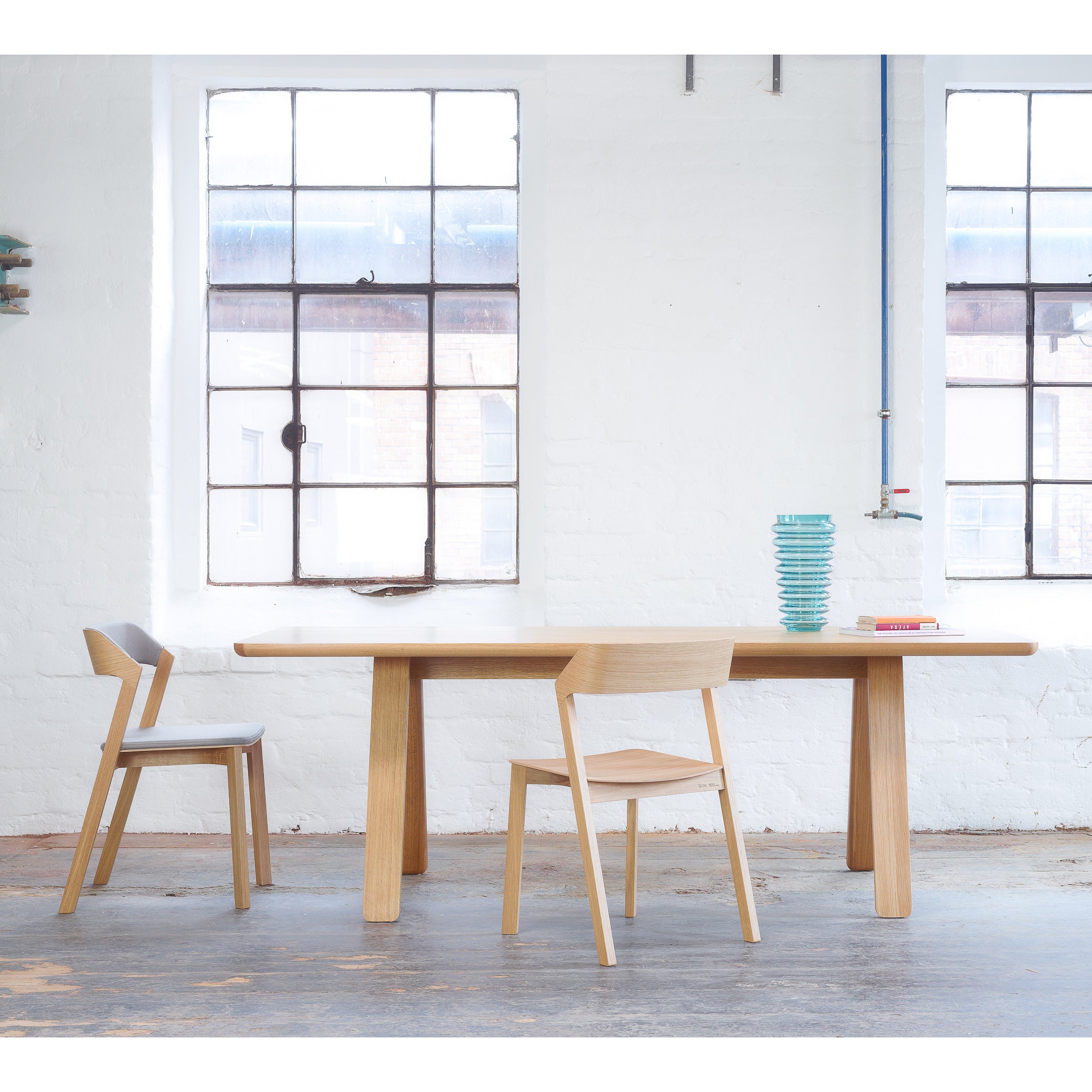 In a minimalist dining room with a concrete floor, the Table Stelvio by TON, crafted from solid wood, is accompanied by two matching chairs. It holds turquoise bowls and books while large industrial windows gently illuminate the space with soft natural light.