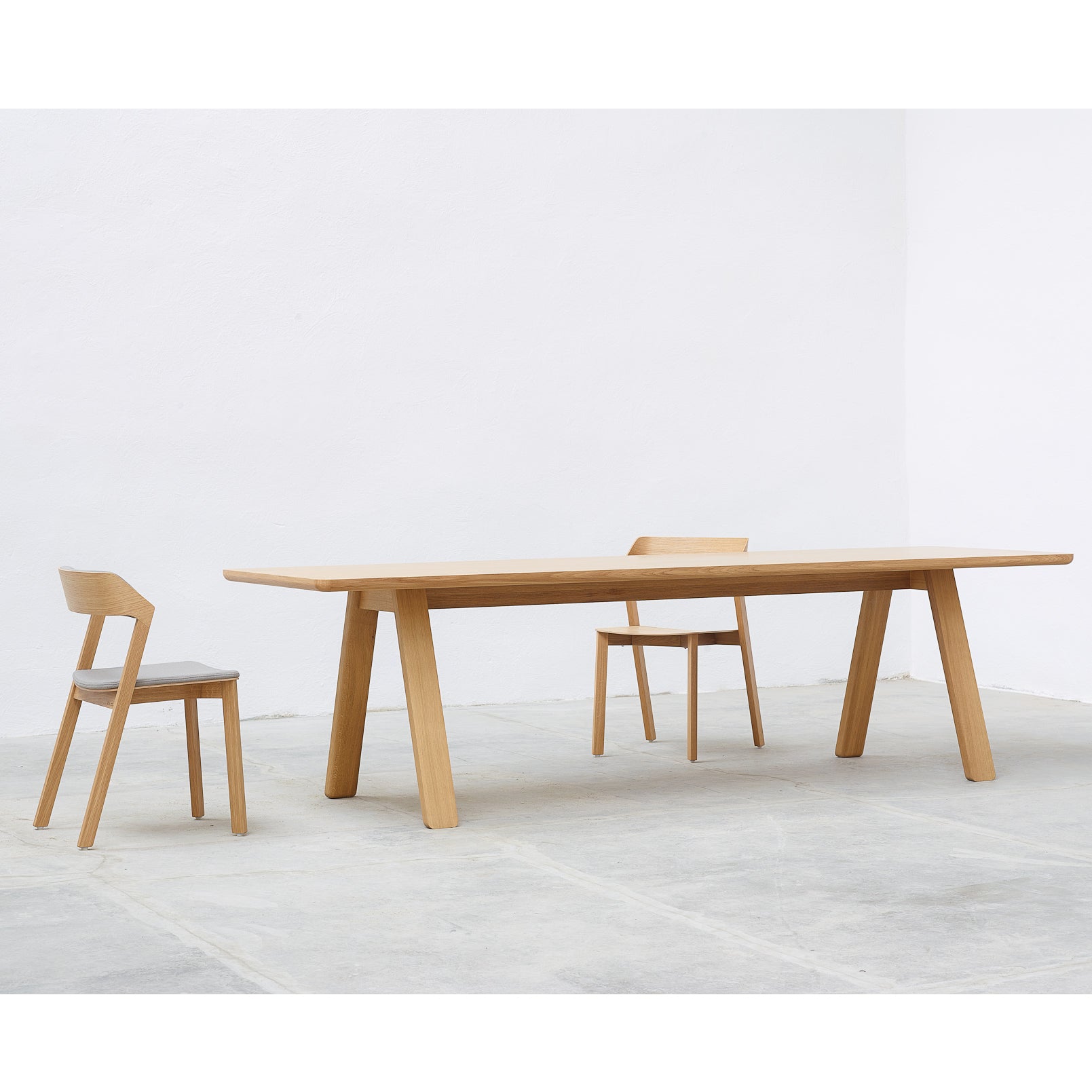 The minimalist Table Stelvio by TON includes a solid wood table with angled legs and two matching chairs, set in a bright room with a light gray floor and white walls, showcasing a clean, timeless design and modern elegance.