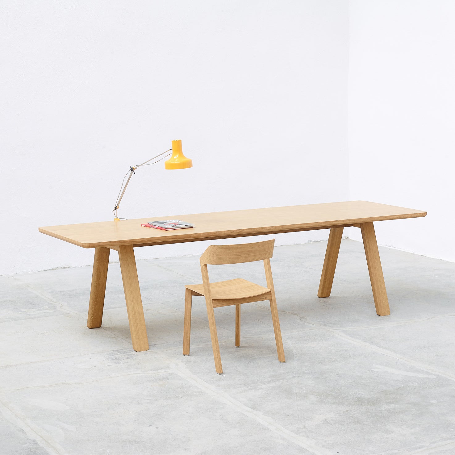 A minimalist room showcases a timeless design with the TON Table Stelvio and a matching chair. A bright yellow desk lamp and two magazines rest on the table. The space, featuring white walls and a light gray floor, exudes a clean, modern atmosphere.