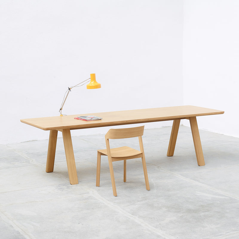 A minimalist room showcases a timeless design with the TON Table Stelvio and a matching chair. A bright yellow desk lamp and two magazines rest on the table. The space, featuring white walls and a light gray floor, exudes a clean, modern atmosphere.