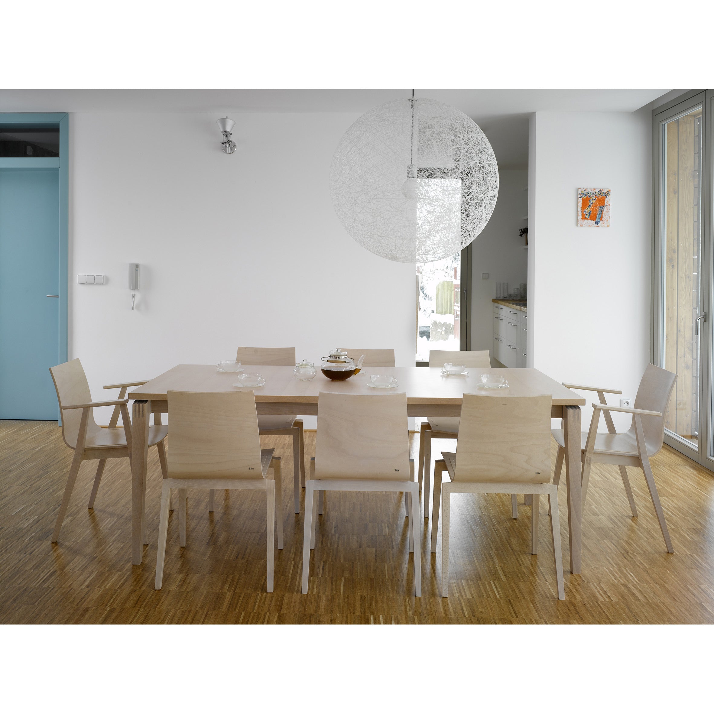 The modern dining room showcases Scandinavian design with a rectangular wooden table and eight TONs Chair Stockholm in pale wood. A large spherical pendant light hangs above, set against a light parquet floor, a blue door to the left, and a small colorful painting on the white wall.
