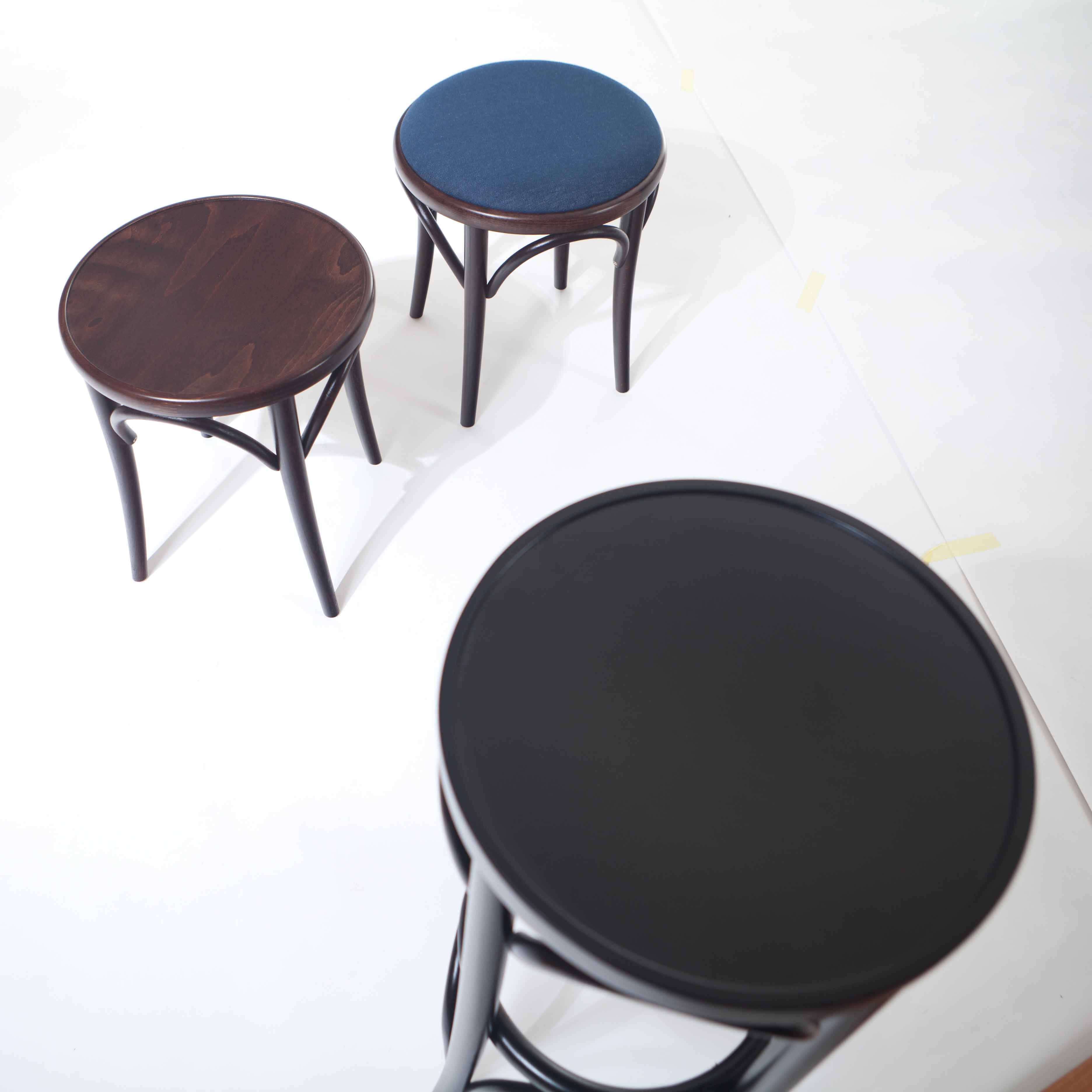 Three round TON Stool 60 Upholstered with beech wood legs are arranged on a light surface: one with a dark wood seat, another with a blue upholstered seat, and the third in black. The slightly elevated angle adds dynamism to the composition.