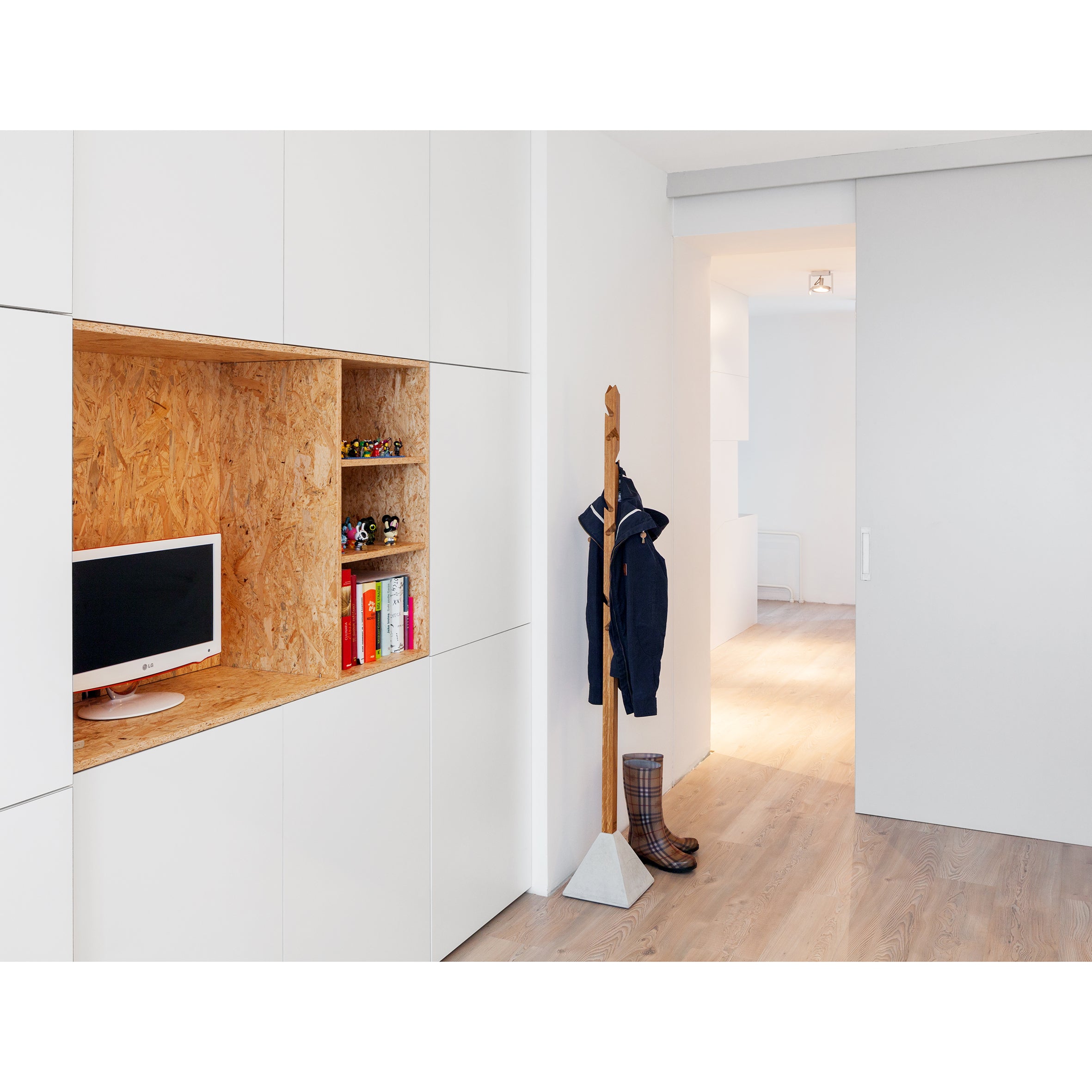 A minimalist room with white walls and light wood flooring features a built-in shelf with chipboard backing holding a monitor and books. Beside it stands the stylish Tee coat rack by TON, made from oak and concrete, while a sliding door opens to reveal a bright hallway.