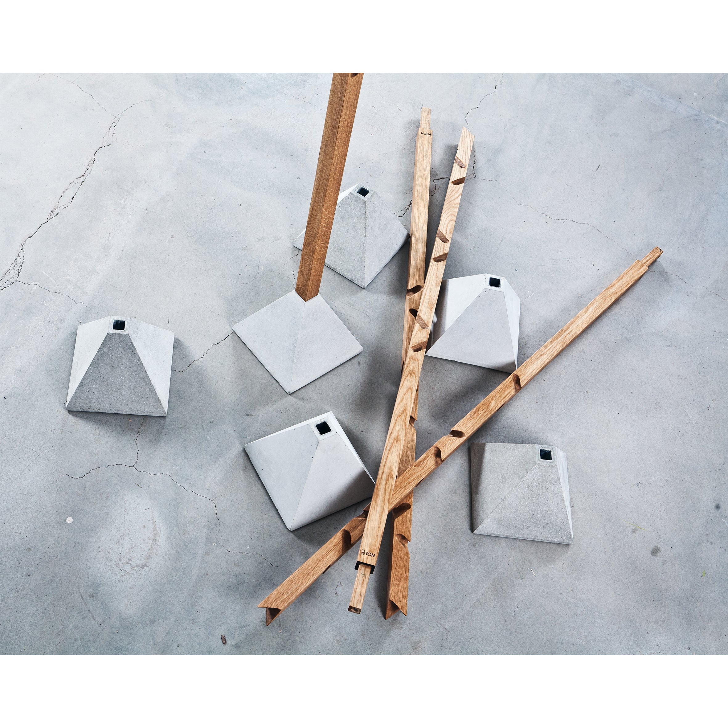 Scattered wooden poles with carved designs rest on a cracked concrete floor beside geometric bases, forming Tee from TON. This minimalist coat hanger cleverly combines a multi-level design with the sturdy elegance of oak and concrete.