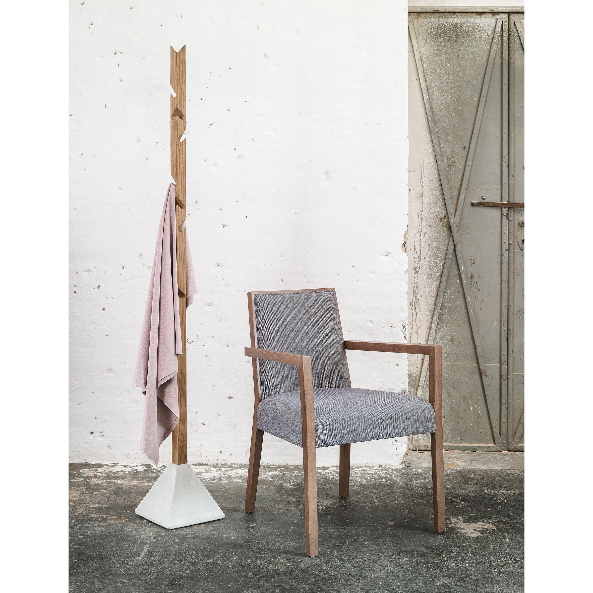 A minimalist room showcases the Tee oak and concrete armchair by TON, upholstered in gray fabric. Nearby, a wooden coat stand stylishly holds light pink fabric. The textured wall and metal door enhance the serene ambiance.