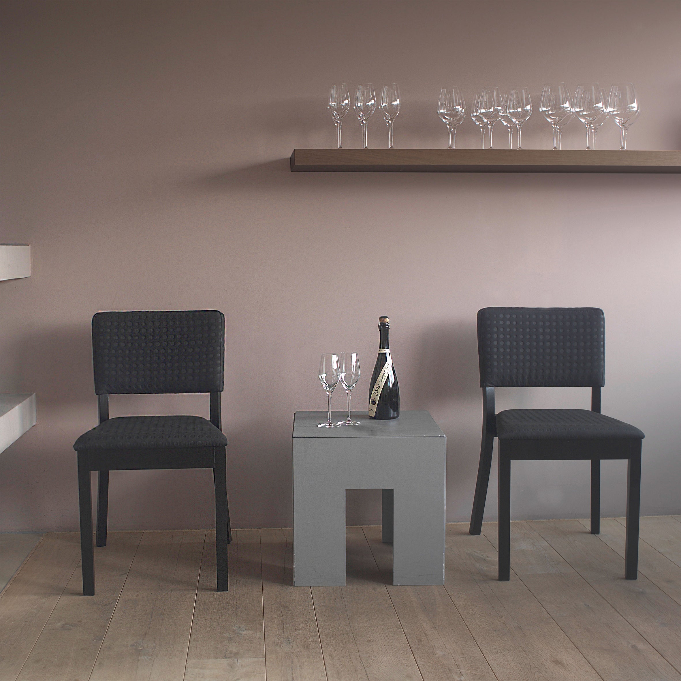 Two black Chair Treviso seats from TON face a small gray table with a champagne bottle and two glasses. Beige walls feature a shelf of empty wine glasses, complementing the wooden flooring.