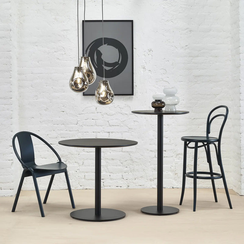 A modern minimalist room features a pedestal table and two black tables of varying heights, including the TON Easy Bistro Table Round. A low chair, high bar stool, pendant lights, a framed abstract print on a white brick wall, and light wood flooring complete the look.