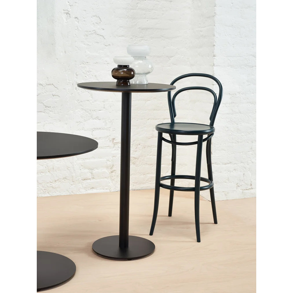 The Easy Bar Table Round by TON stands tall with a steel base and black finish, paired with a sleek black stool against a white brick wall. Two small vases—one white, one dark brown—top the table. Light wooden flooring contrasts beautifully with this chic setup.