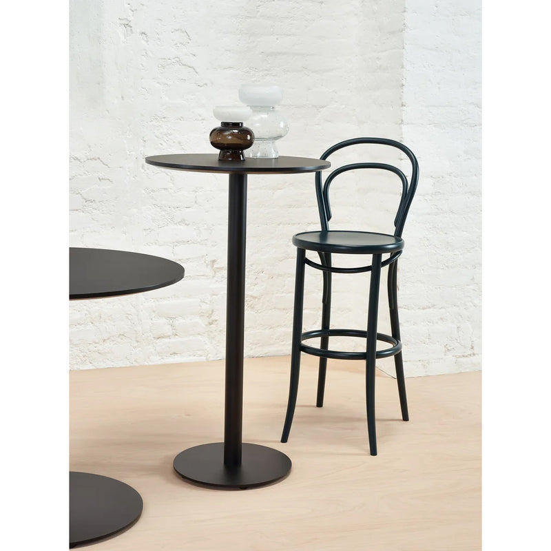 The Easy Bar Table Round by TON stands tall with a steel base and black finish, paired with a sleek black stool against a white brick wall. Two small vases—one white, one dark brown—top the table. Light wooden flooring contrasts beautifully with this chic setup.