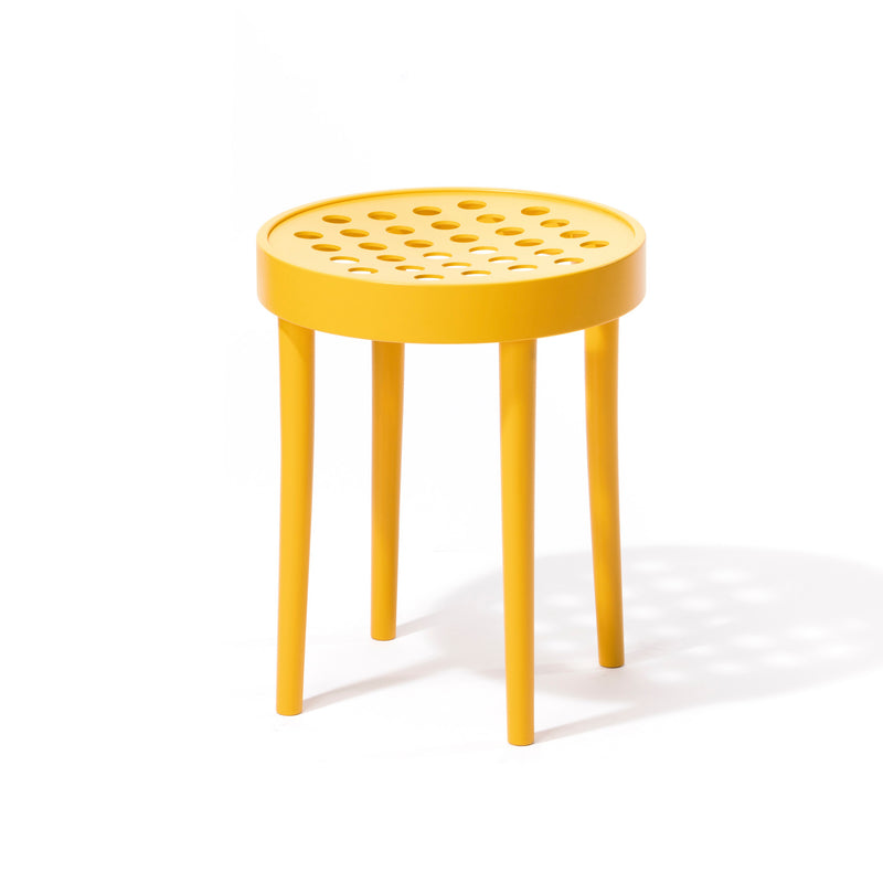 The Stool 822 by TON features a round yellow seat with a pattern of small holes, showcasing Scandinavian minimalism. Designed in the style of Claesson Koivisto Rune, it has four tapered legs and softly casts shadows on a white background.