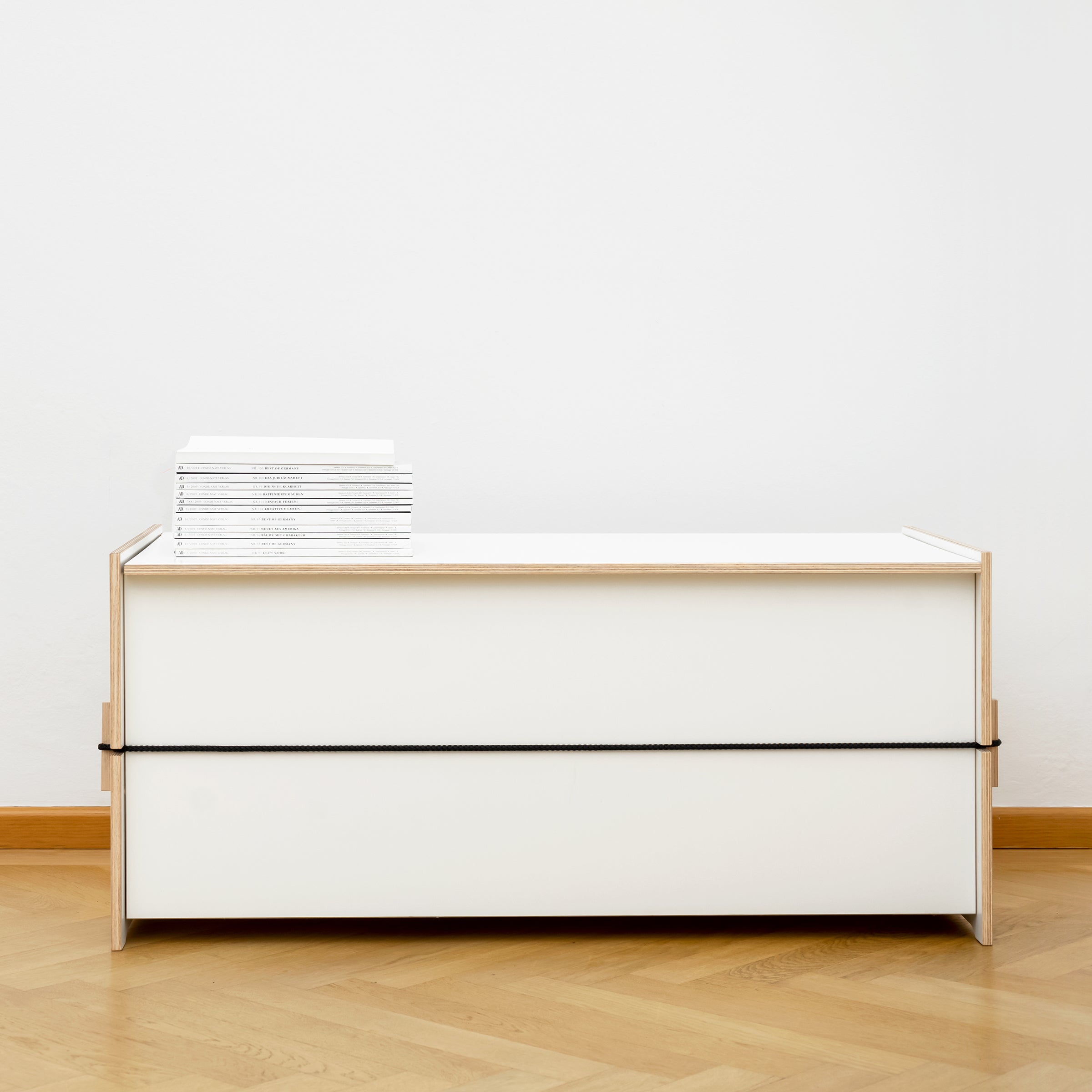 Against a white wall and wooden floor, the modern minimalist Trude unit by Moormann features a light wooden frame. Its spacious surface holds neatly stacked white books, making it perfect for storing bed linen.