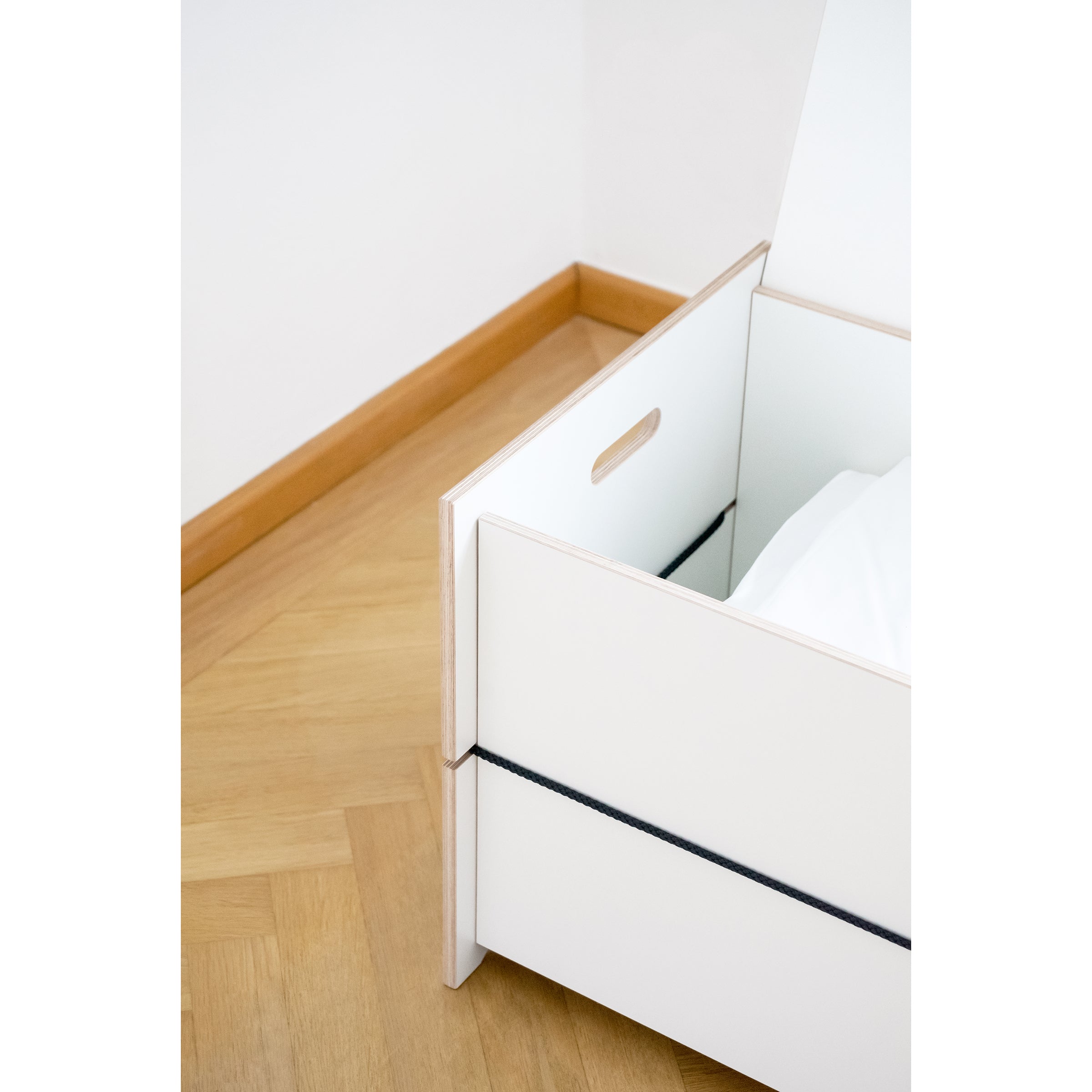 A white Moormann Trude storage box with a cut-out handle rests on the parquet floor, ideal for storing bed linens. Partially open, it shows a fabric lining inside. The walls are painted white with a light brown baseboard in the corner.
