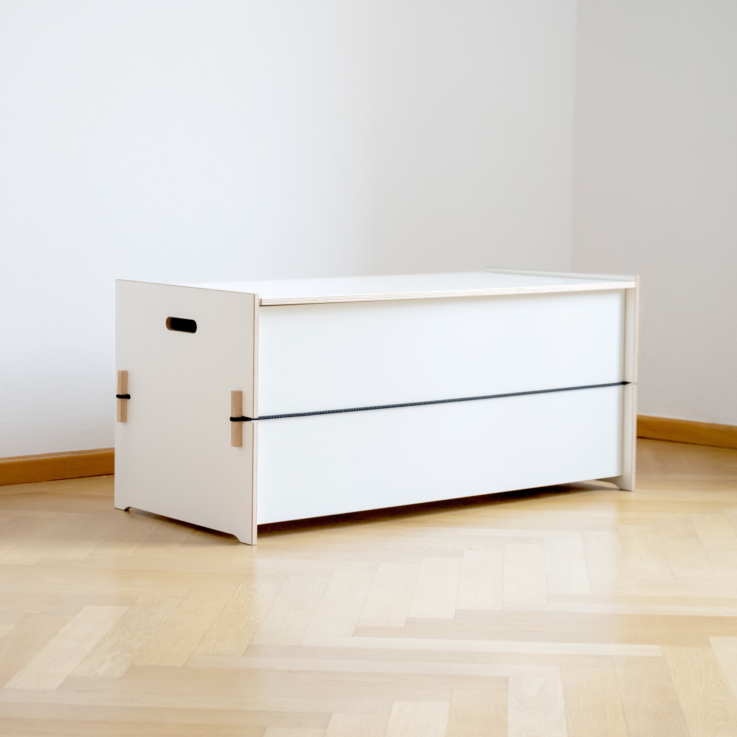 The Trude by Moormann is a minimalist white storage box with a sleek black band and side handles. Ideal for bed linen storage, it complements a room with white walls and a light wooden floor.