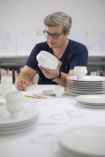 A person wearing glasses examines a white, Augarten Shortcut - Sale bowl while sketching on paper. Various stacked white ceramic dishes occupy a table surrounded by sketches, creating a creative scene with shelves filled with books and more sketches in the background.