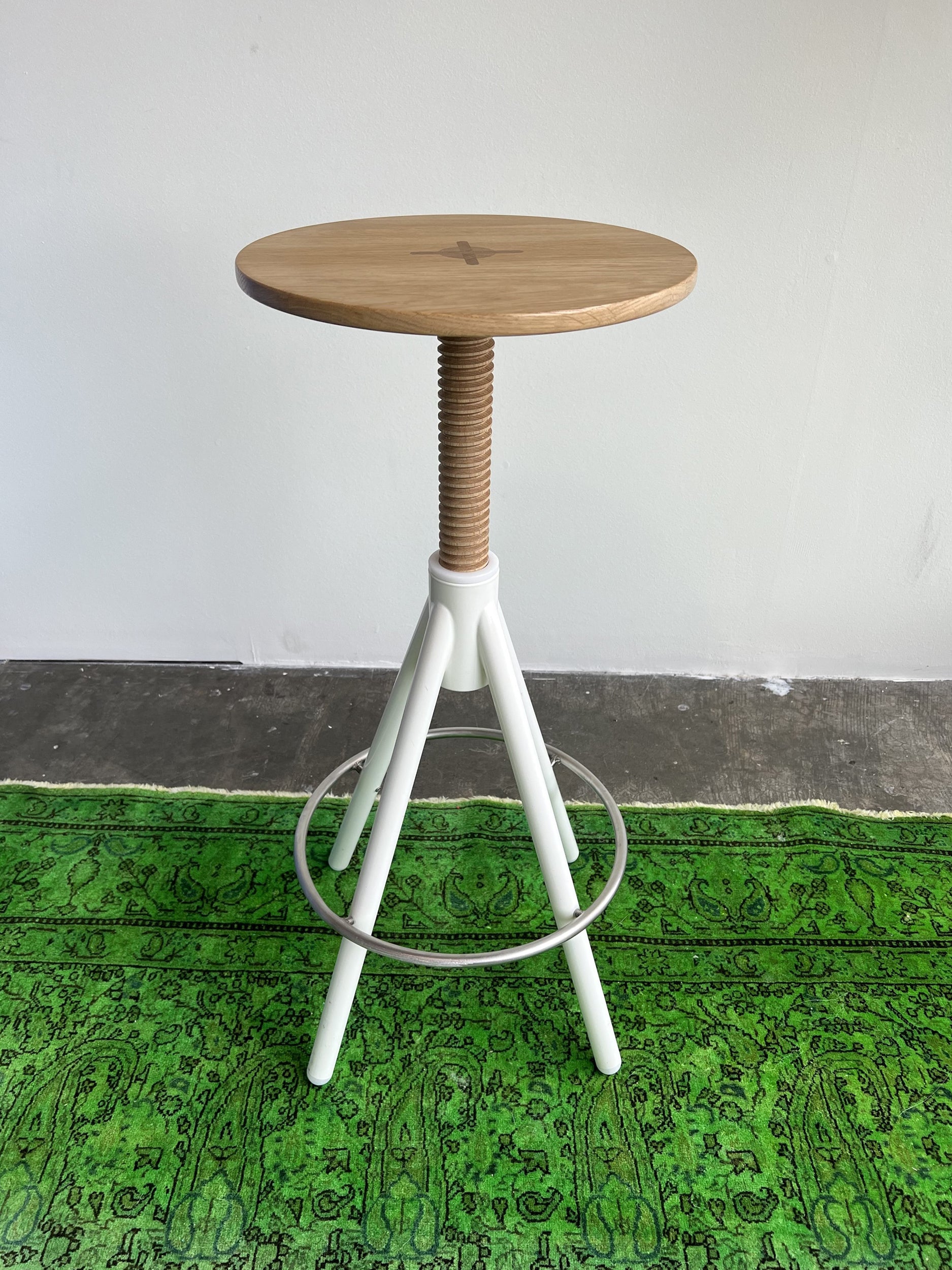 The Thread Family High Stool by Coordination Berlin features a walnut wooden seat with a white adjustable screw stem and tripod legs. This elegant, height-adjustable stool complements a green patterned rug against a pristine white wall.