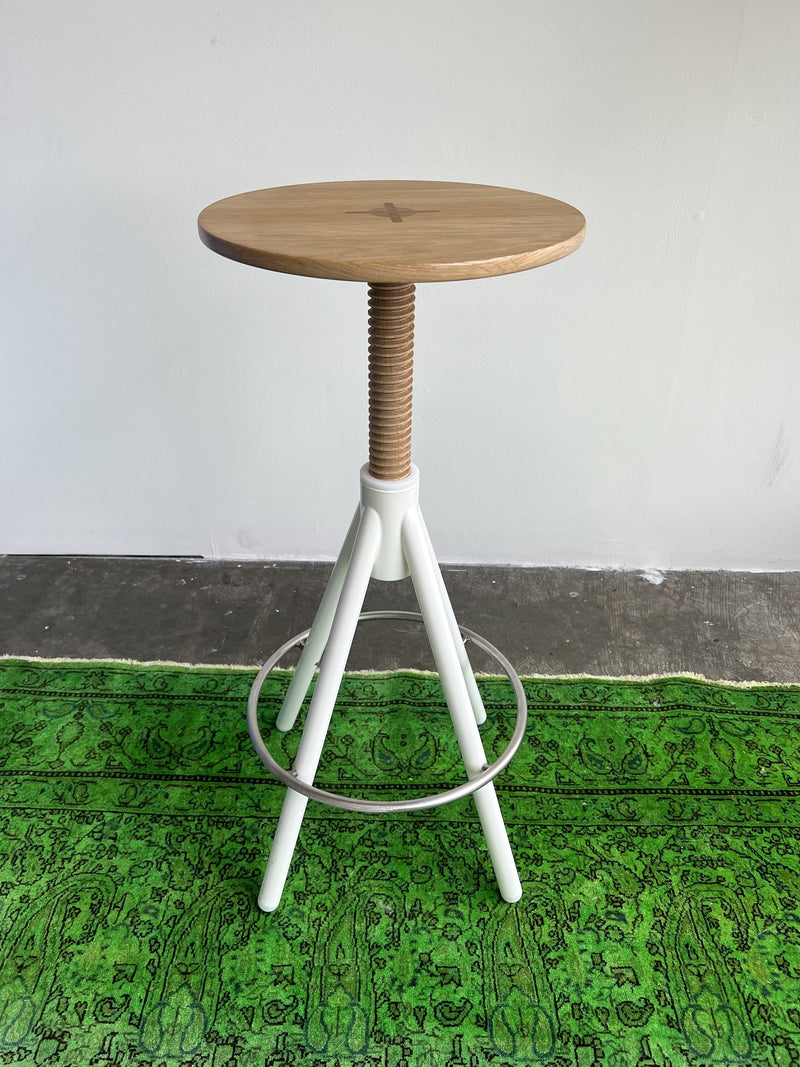 The Thread Family High Stool by Coordination Berlin features a walnut wooden seat with a white adjustable screw stem and tripod legs. This elegant, height-adjustable stool complements a green patterned rug against a pristine white wall.