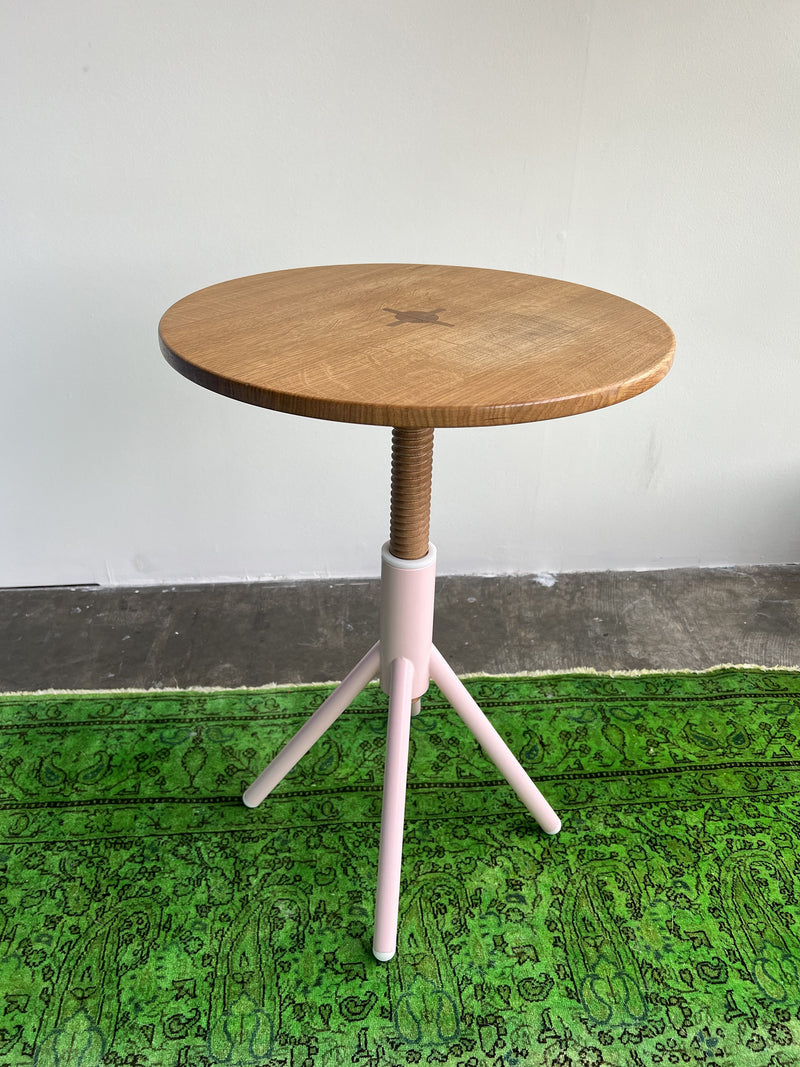 The Thread Family Bistro Table by Coordination Berlin features a screw-adjustable round wooden design with a light oak top and white welded steel three-leg base, set on intricately patterned textured green carpet against a plain white wall.