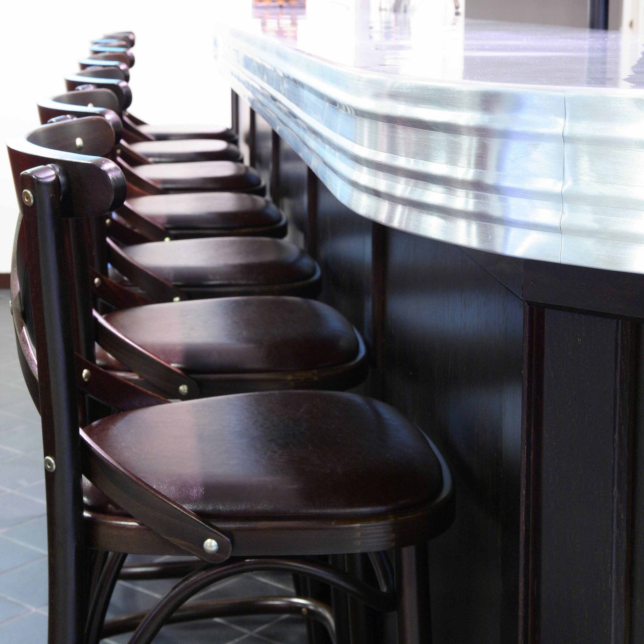 TONs Barstool 150, crafted from rich dark wood, lines the sleek metal countertop in a bar or restaurant setting. The spotless tile floor enhances the inviting and timeless ambiance of this refined space.