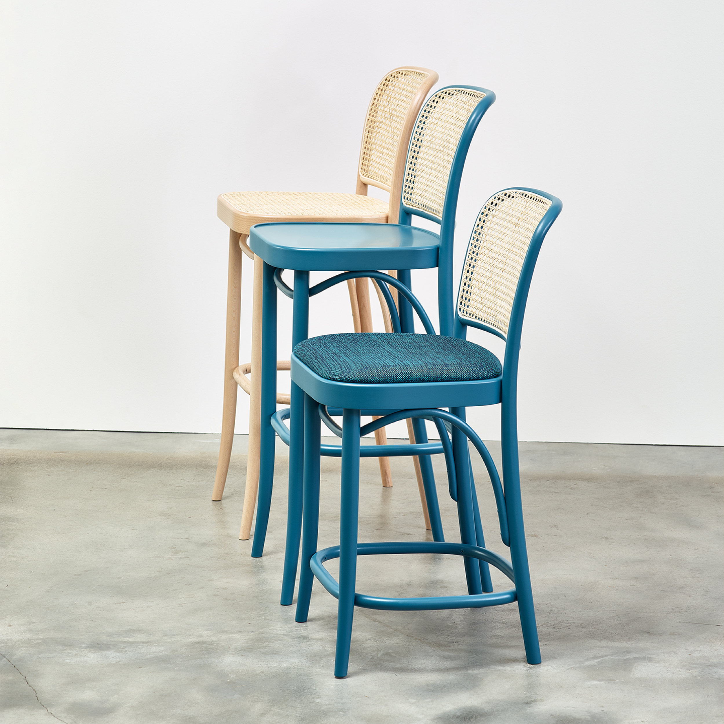 Three TON Barstool 811 models are lined up. The first showcases light beech wood with a cane mesh backrest, the second has a blue seat and woven pattern back, and the third is all blue. They elegantly contrast against a gray concrete floor and white wall.