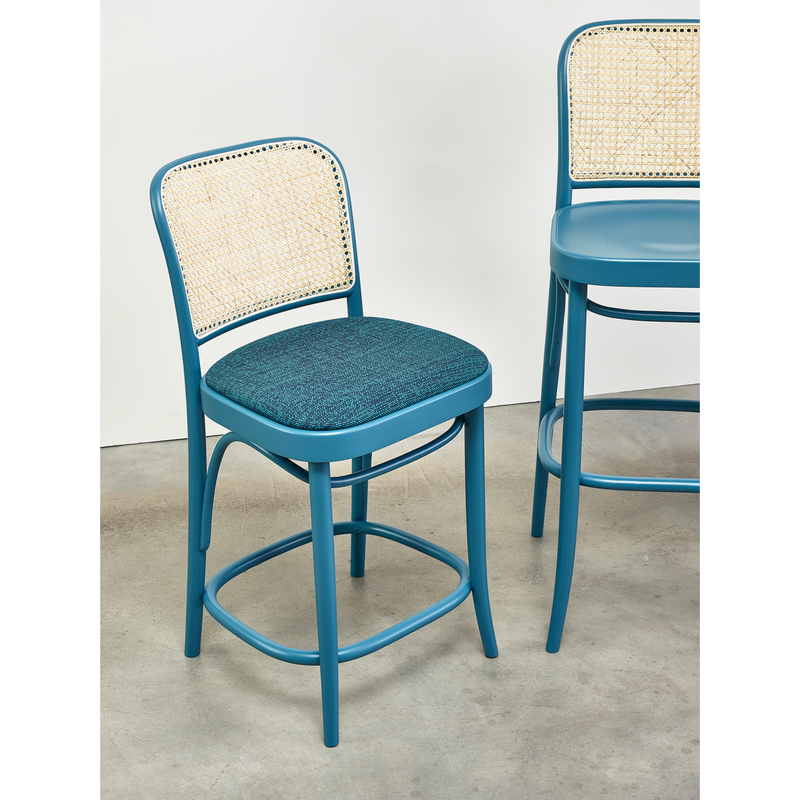 The TON Barstool 811 - Upholstered, with a blue cushioned seat and woven cane mesh backrest, stands on a concrete floor. Another similar stool is partially visible. Both boast a sleek design with curved beech wood legs.