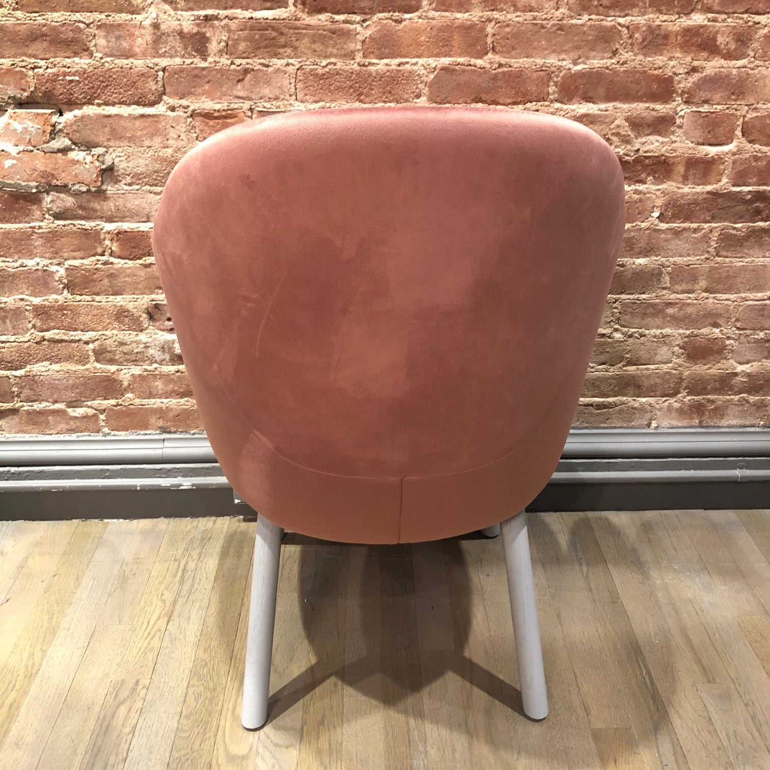 The Armchair Albu by TON, a high-quality upholstered pink chair with a rounded back and ergonomic design, is positioned against a brick wall. It features four light gray legs and rests gracefully on a wooden floor.