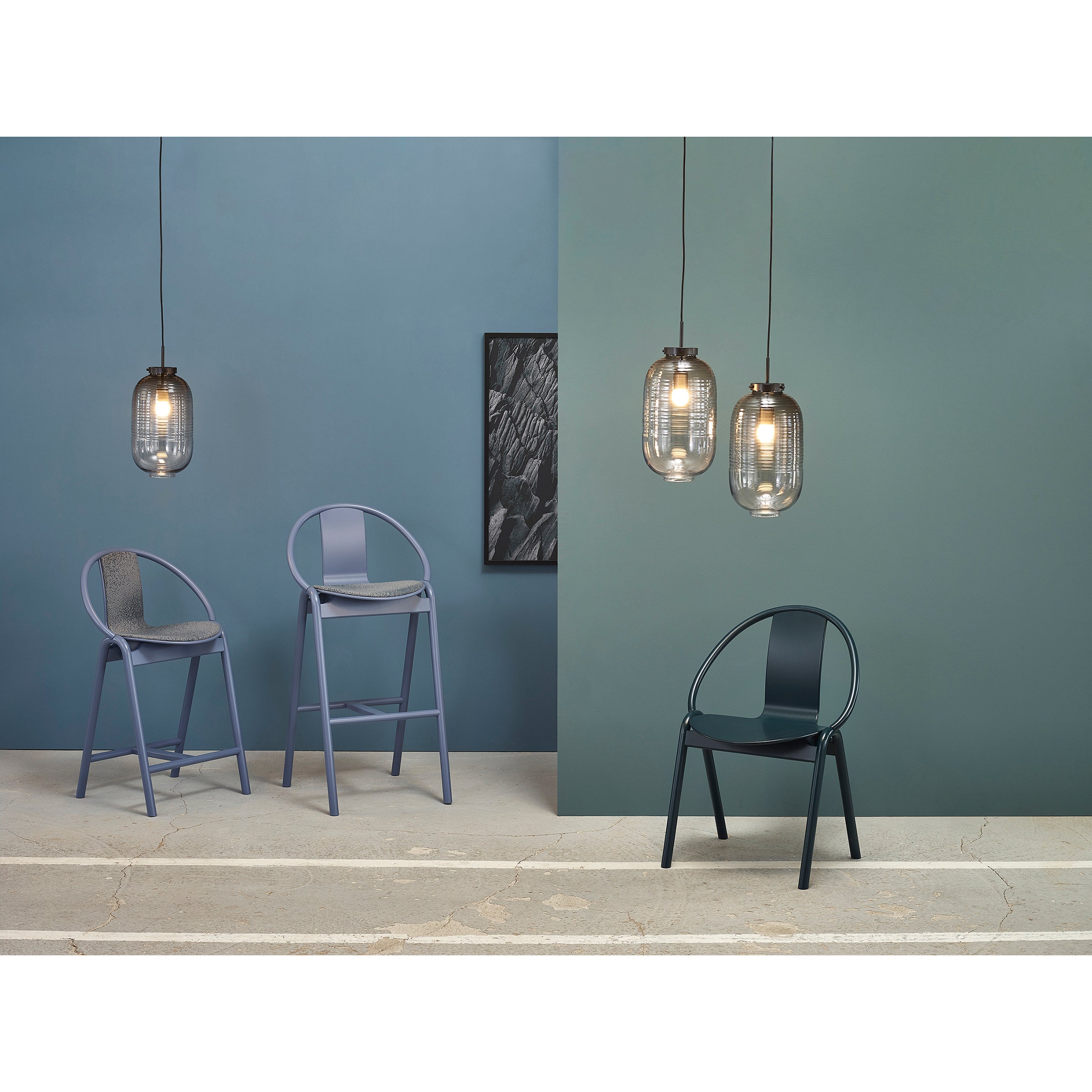 A modern interior features two light gray bentwood Barstool Again Upholstered by TON with curved backs, a dark gray chair, three hanging glass pendant lights, and a framed black-and-white artwork on a blue-gray wall. The floor has textured concrete.