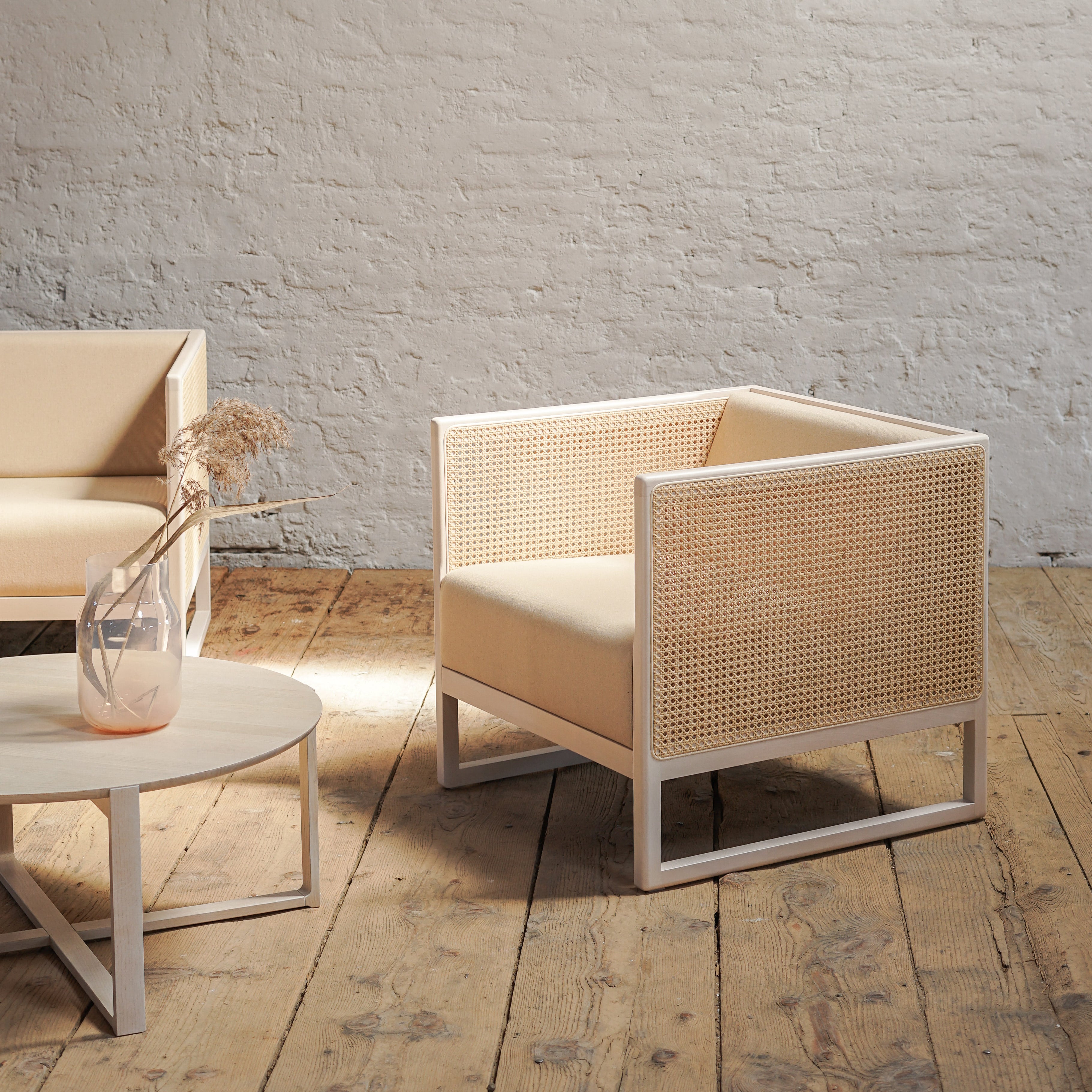 A minimalist room showcases the cream-colored Armchair Casablanca by TON, featuring cane weave armrests paired with a matching two-seater sofa. A round wooden table holding dried flowers sits on a rustic wooden floor against a white brick wall, softly illuminated by natural light.