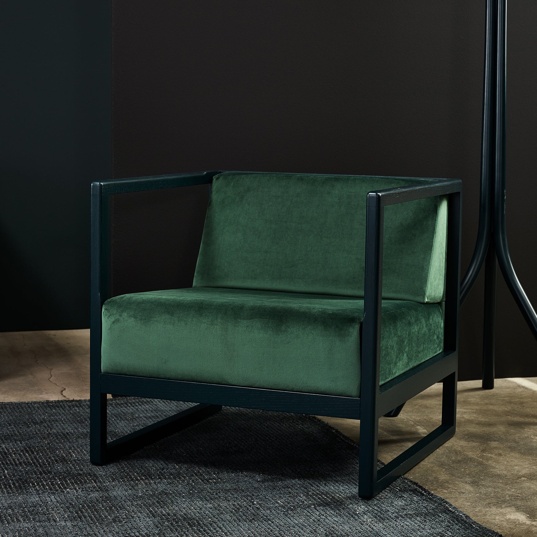 The Armchair Casablanca by TON, with dark green velvet cushions and a black metal frame, rests on a dark rug. A black wall serves as the background while a floor lamp peeks beside it. Elegantly crafted cane weave armrests enhance this sophisticated setup.
