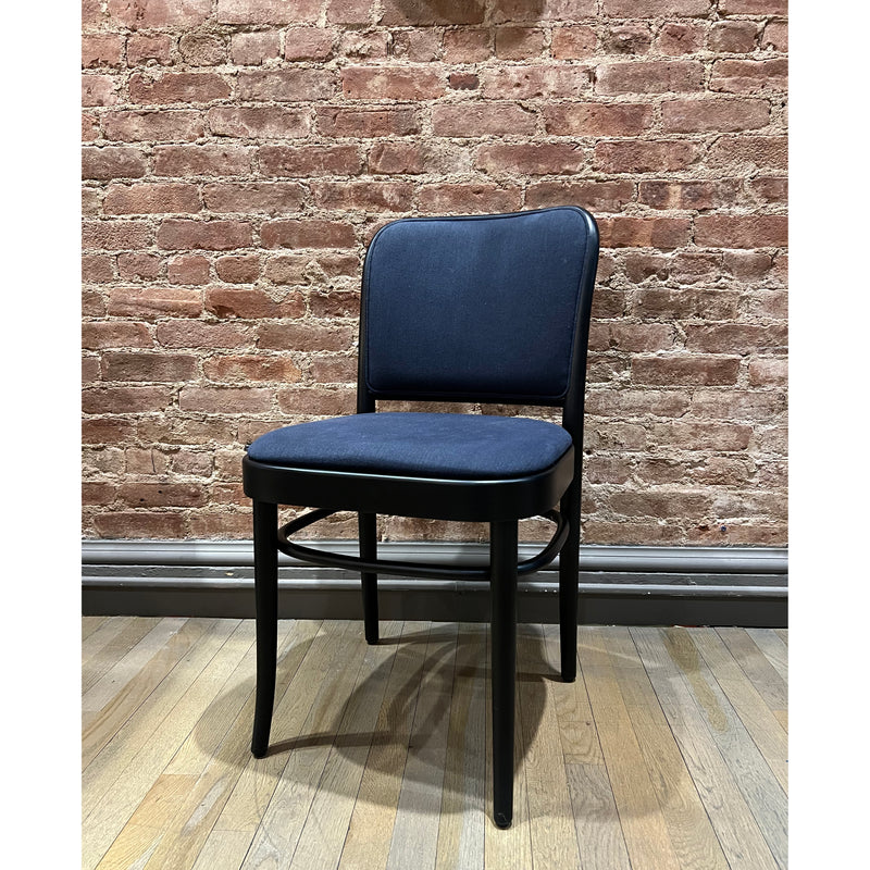The Chair 811 by TON exudes Art Nouveau elegance with its black wooden frame and padded navy blue seat and backrest, beautifully contrasted against a polished wooden floor and exposed red brick wall.