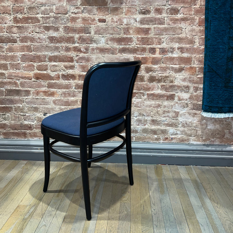 A Chair 811 by TON, upholstered in blue and reminiscent of Josef Hoffmanns design, sits elegantly on a wooden floor against a brick wall. The black frame contrasts beautifully, with a hint of blue curtain enhancing its Art Nouveau charm.