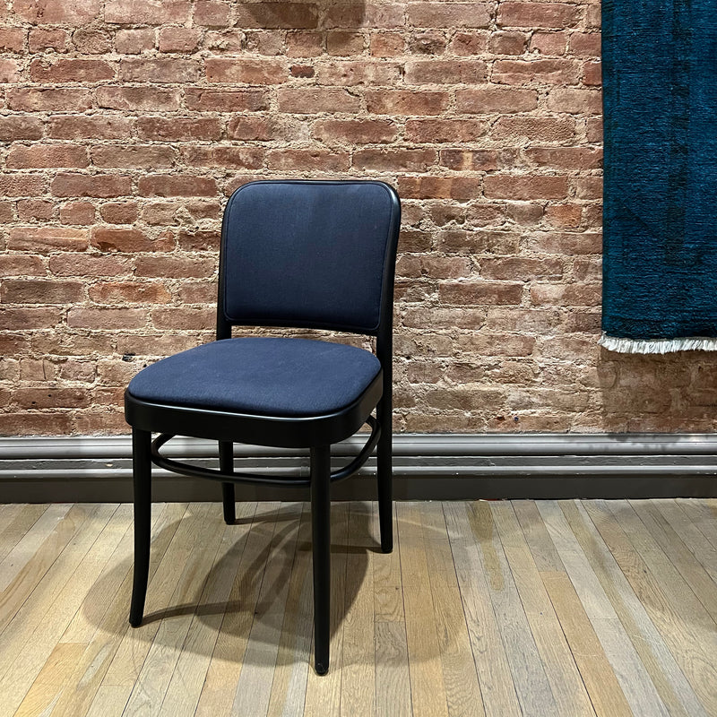 The Chair 811 by TON, with dark blue upholstery and a black frame evoking Art Nouveau elegance, sits on a wooden floor against a textured brick wall. A dark blue curtain with white fringe partially visible on the right enhances the sophisticated ambiance.
