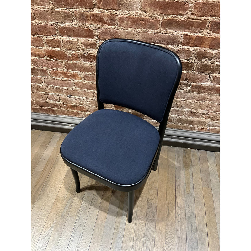 The Chair 811 - Sale from TON, featuring a black frame and dark blue cushioned seat and backrest, sits on a wooden floor. An exposed red brick wall in the background enhances its Art Nouveau charm with a vintage appeal reminiscent of Josef Hoffmann design.