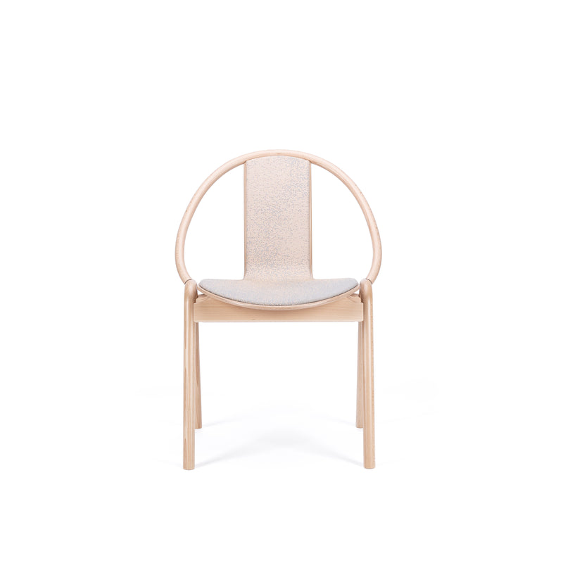 The Chair Again Upholstered by TON is a minimalist design crafted from solid beech wood, featuring a smooth circular backrest and cushioned seat. Its modern and elegant structure boasts light-colored wood, enhancing its sophisticated look against white backgrounds.
