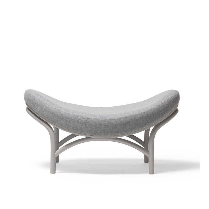 The Ottoman Chips by TON is a stylish, gray, curved ottoman with a minimalist design that complements the Chips lounge chair. It features a smooth, cushioned seat for comfort and light wooden legs bent manually into an intersecting arch pattern for an airy appearance against a white backdrop.