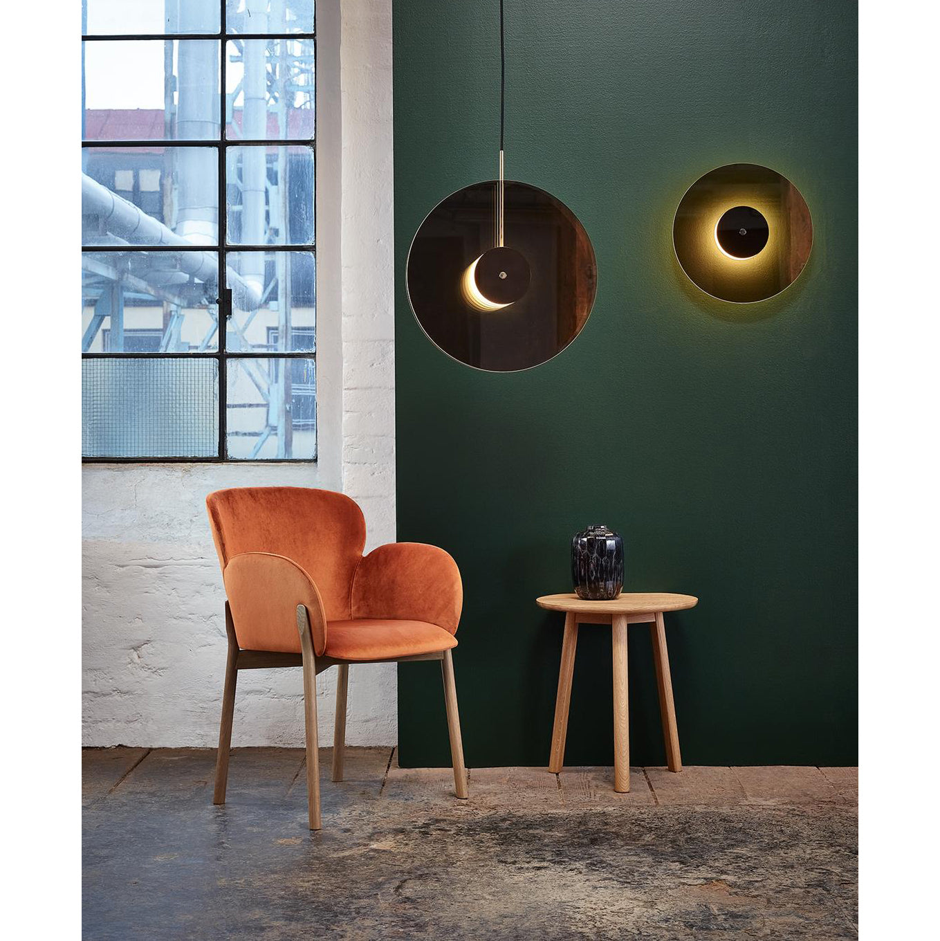 A modern room features an Armchair Ginger by TON in plush orange, next to a small table made with the TON wood-bending technique. A dark vase completes the setup, while two circular glowing wall lights enhance the green wall beside an industrial-style window filtering soft daylight.