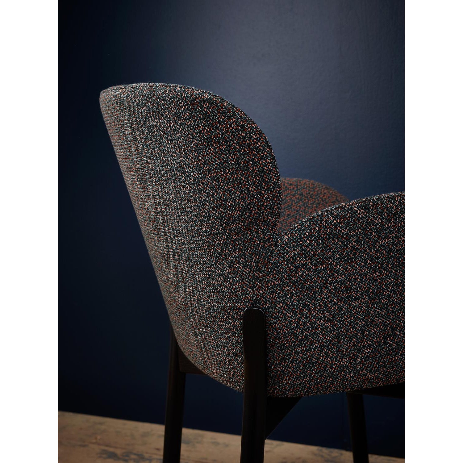 A close-up of the Armchair Ginger by TON reveals a rounded backrest against a dark blue wall. The textured, multicolored fabric and black legs highlight TONs wood-bending technique, while the minimalist setting enhances its modern, contemporary design.