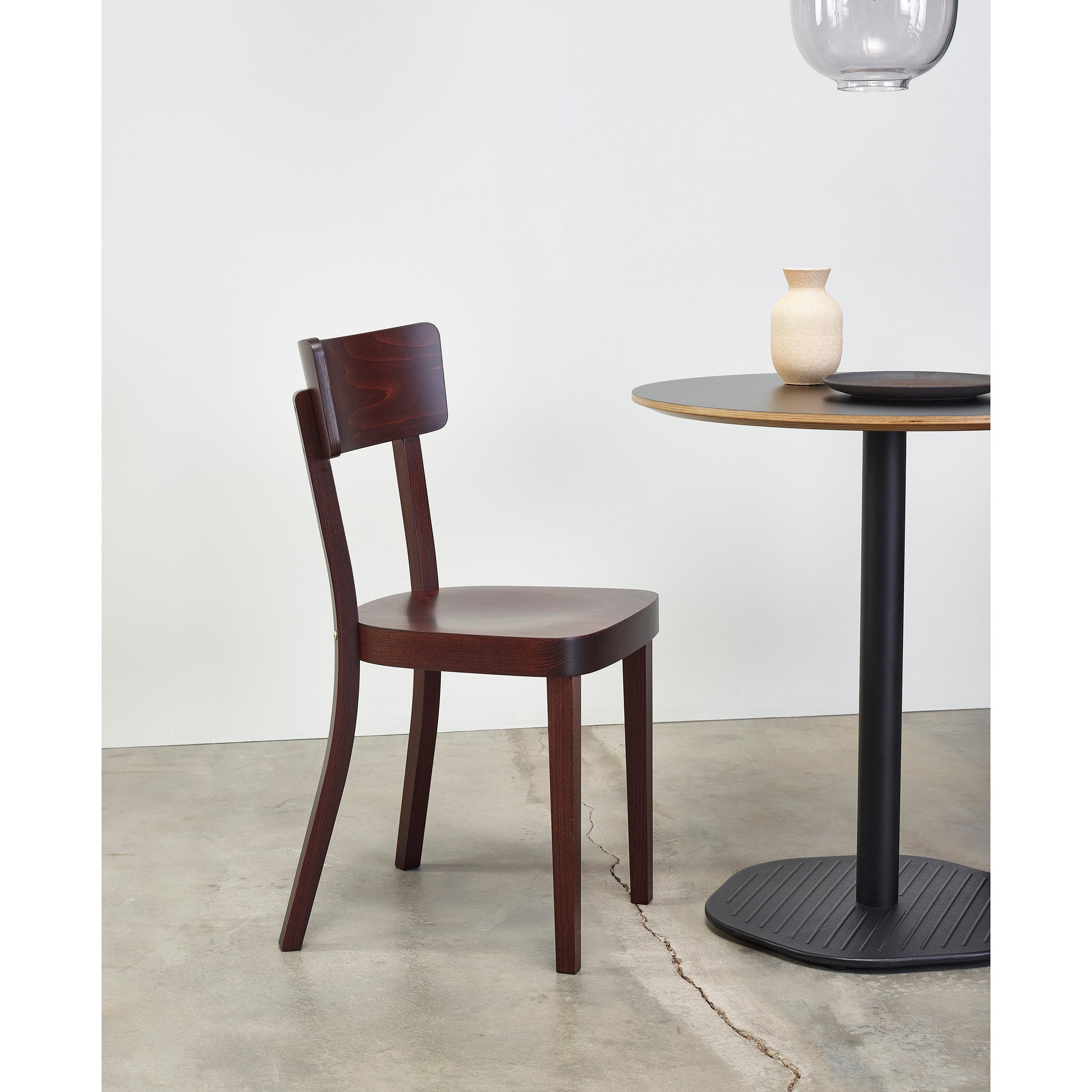 A TON Chair Ideal in dark wood with an upholstered seat sits beside a round table with a black base. On the table, theres a small beige vase and a black dish, forming a perfect dining scene against a plain white wall on a concrete floor.