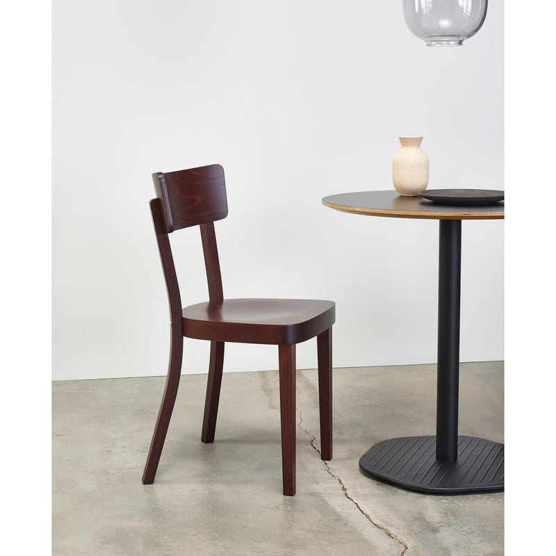 A TON Chair Ideal in dark wood with an upholstered seat sits beside a round table with a black base. On the table, theres a small beige vase and a black dish, forming a perfect dining scene against a plain white wall on a concrete floor.