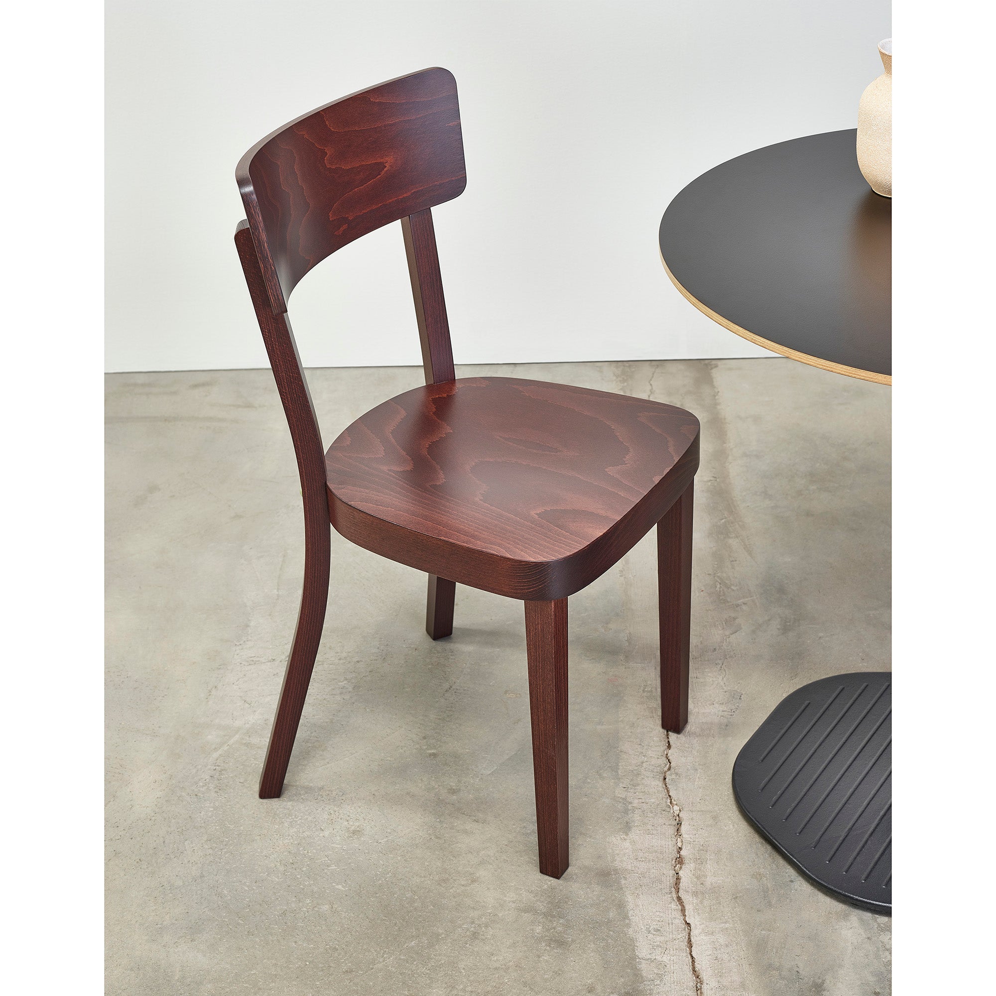 The Chair Ideal by TON, made from beech with a curved backrest and dark finish, sits next to a round dark table on a concrete floor. Perfect for the hospitality industry.