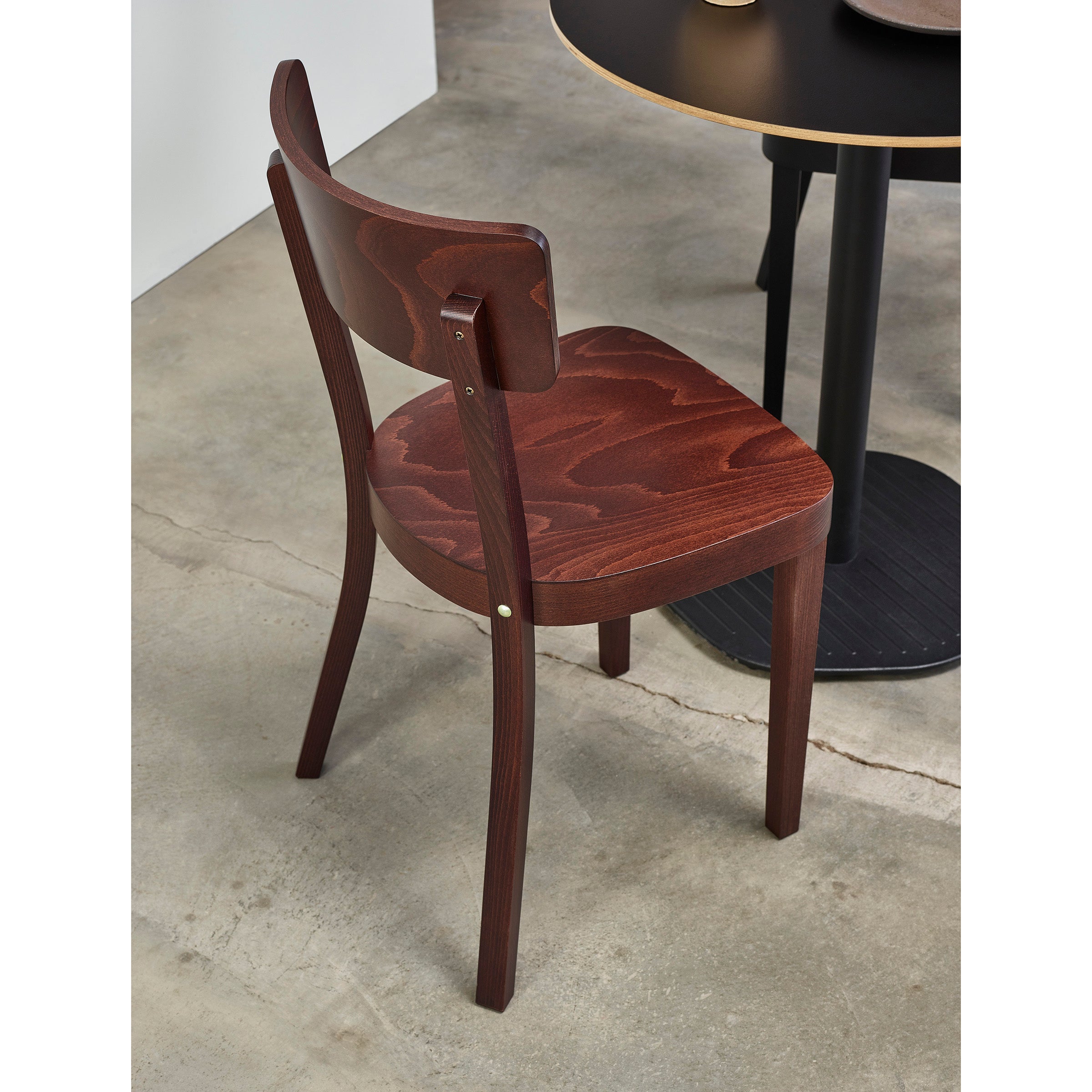The Chair Ideal by TON features an upholstered seat and curved backrest in a rich beech finish, showcasing beautiful grain patterns. This stylish wooden chair complements a round black table perfectly against the polished concrete floor, ideal for those who value comfort and elegance.