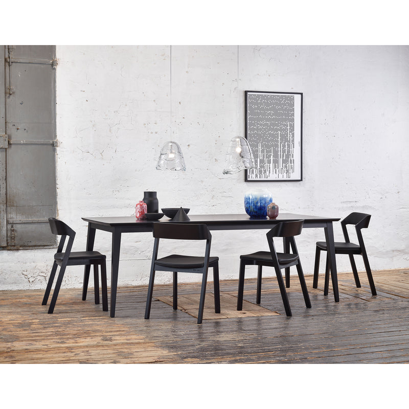 A modern dining room features the Jylland Table by TON, a long black extendable piece with five matching Scandinavian chairs. Two transparent pendant lights illuminate vases and bowls, while a textured white brick wall and abstract artwork enhance the minimalist decor.
