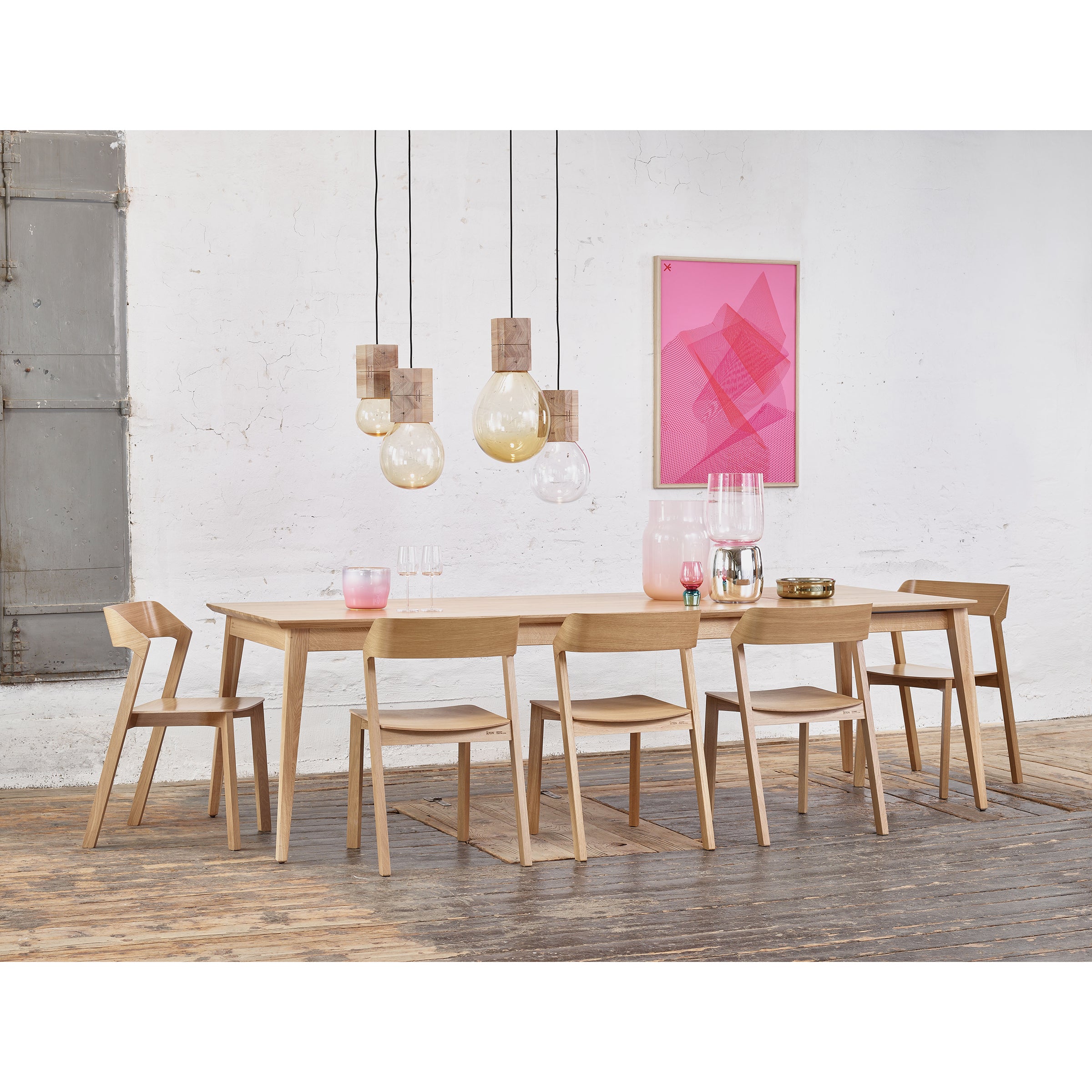 A modern dining room showcases a solid wood Jylland Table by TON with six chairs. Pendant lights illuminate a pink abstract artwork on the pristine white wall, while the wooden floor and a partially visible rustic metal door enhance the Scandinavian design charm.